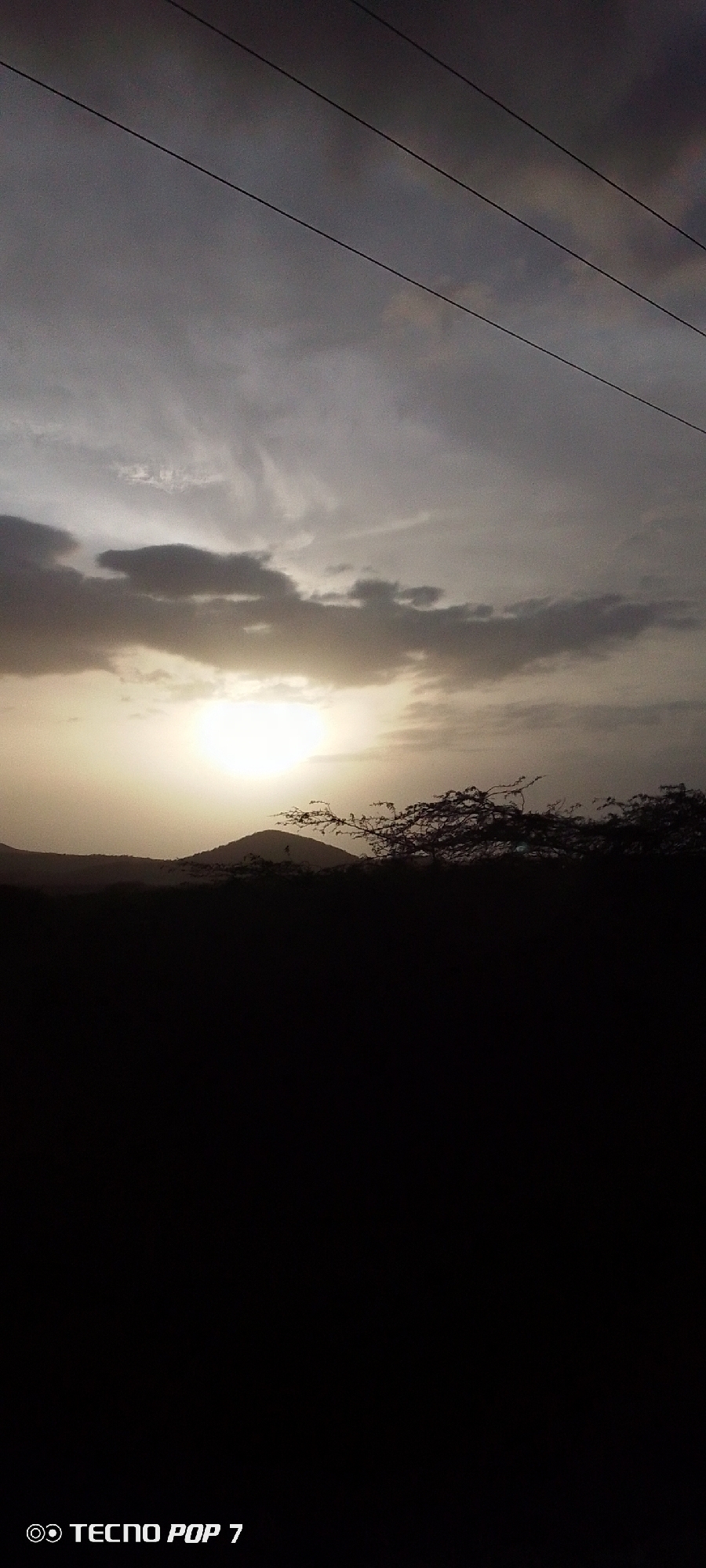 A silhouette image shows a landscape under a cloudy sky at dusk or dawn. The sun is low on the horizon, partially obscured by clouds, casting a bright glow. A dark landmass with rolling hills and vegetation is visible in the foreground. Three parallel power lines are strung across the top portion of the frame. The photo appears to be taken with a "TECNO POP 7" phone. There is no evidence to confirm the location is in Altagracia, Venezuela. No people are present.