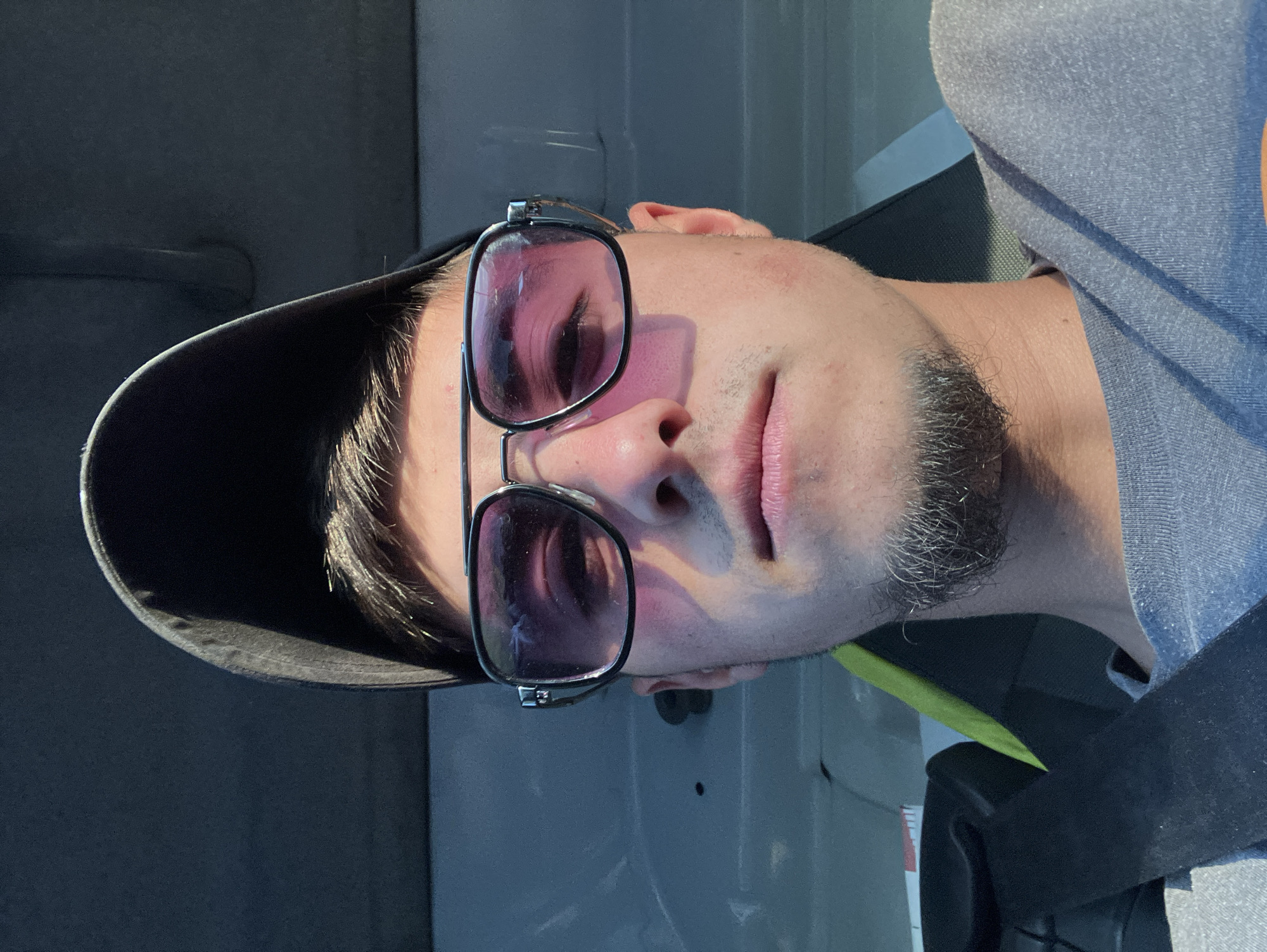 A man is facing the camera, wearing a black baseball cap and sunglasses with a pink tint. He has a short beard and is wearing a gray shirt and a black seatbelt. The background appears to be the interior of a vehicle. There is no information provided to confirm the location of the city of Komuniga, Bulgaria.