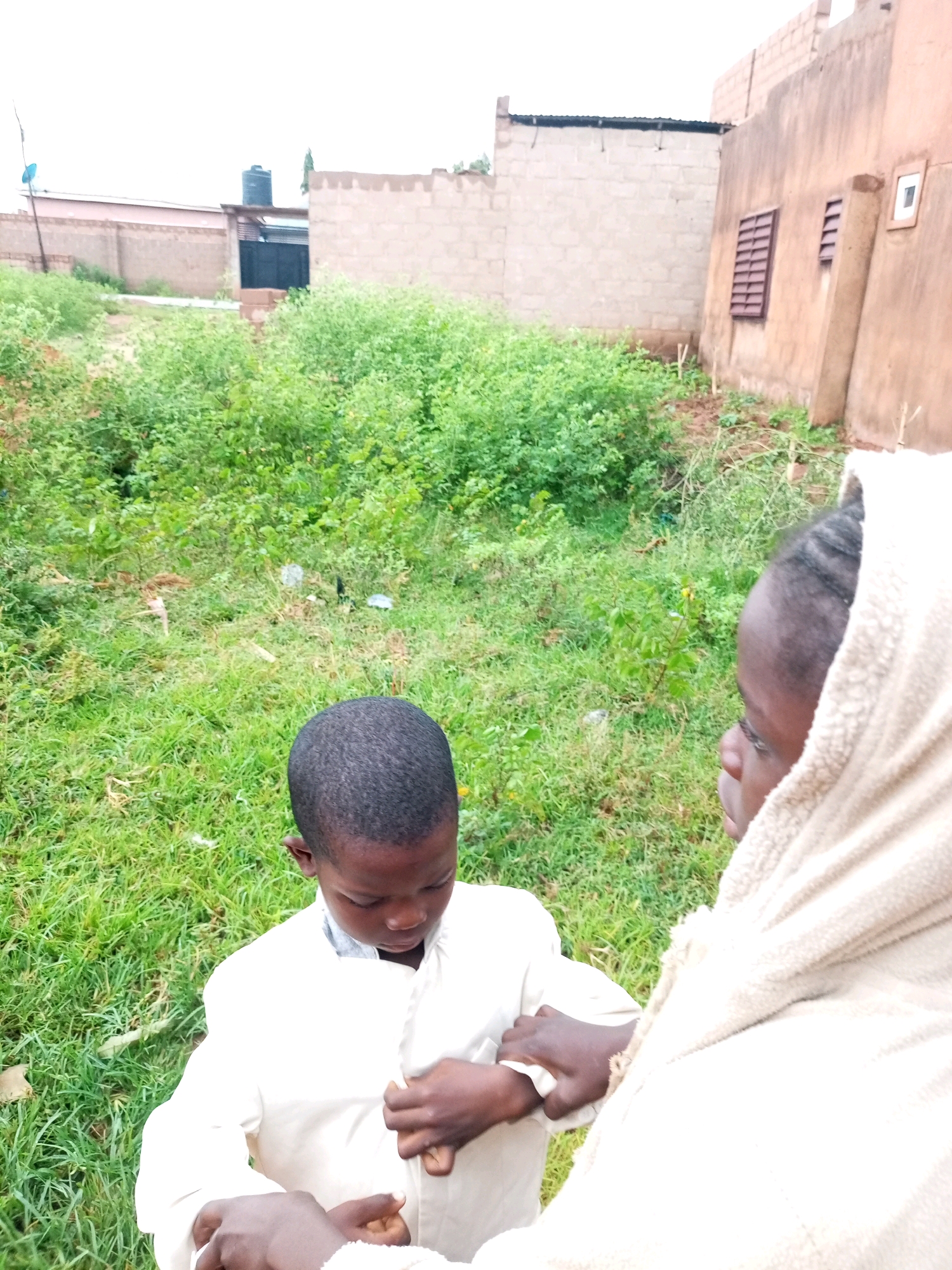 In a partially constructed area of Tempure, Nigeria, two children are engaged in an activity outdoors. The scene is characterized by overgrown vegetation and unfinished buildings. One child, a boy wearing a white shirt, is the focus of attention as his garment is being buttoned by another individual, whose face is partially obscured by a light-colored hooded garment. The backdrop includes walls constructed of concrete blocks, one with a barred window and another with a small white-framed opening. A water tank is visible on a rooftop in the distance. The ground is covered in grass and scattered debris. The lighting suggests an overcast day.