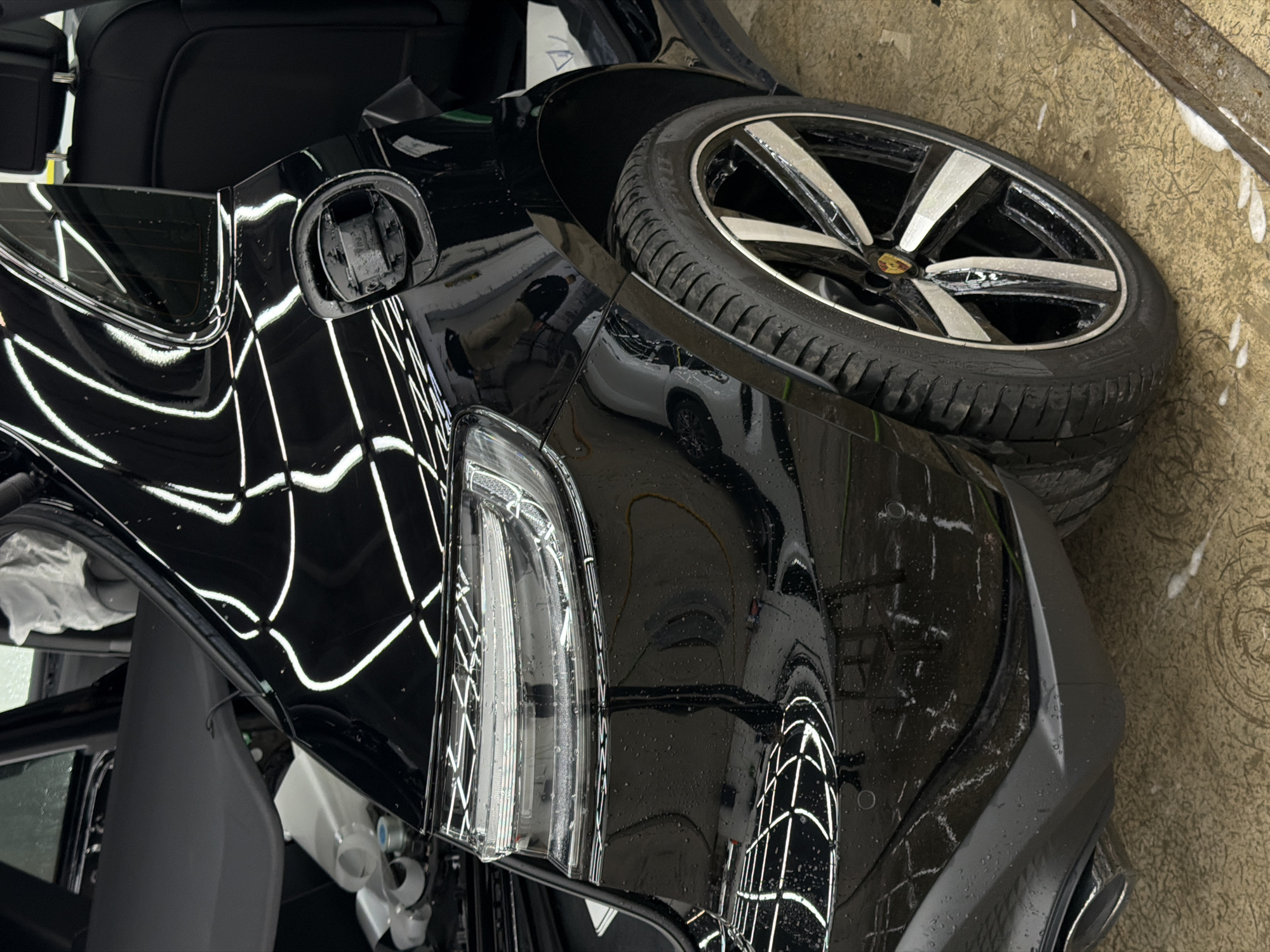 The image shows a close-up of the rear of a black Porsche SUV, likely in a garage or car wash. The car appears to be wet, and light reflects off its surface in a grid-like pattern. The rear wheel is visible, with a silver and black rim featuring the Porsche emblem. The rear taillight is also prominently displayed. The fuel filler door is open. The environment appears to be a concrete floor with water droplets or residue, suggesting the car is undergoing cleaning or detailing. There is no verifiable information in the provided image to determine if the location is Krasnoyarsk, Russia.
