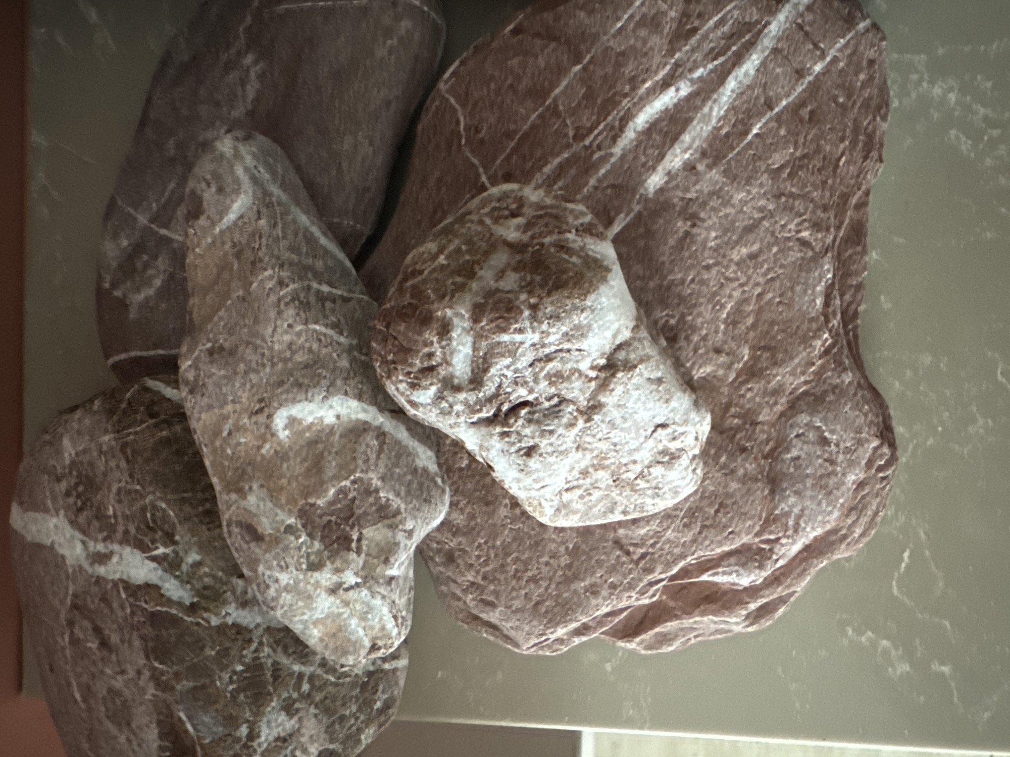 The image shows a collection of rocks on a speckled, light-colored countertop. The rocks are varied in size and color, with dominant earthy tones of brown and red, interspersed with white veining. The surface they rest on appears to be a polished stone or laminate countertop with subtle swirling patterns. The lighting suggests an indoor environment, possibly a home or a shop. There are no people or animals present in the image, and no discernible actions or interactions are taking place. The scene is static and focused solely on the arrangement of these natural stones. The image does not provide any information to suggest it is located in Fethiye, Turkey.