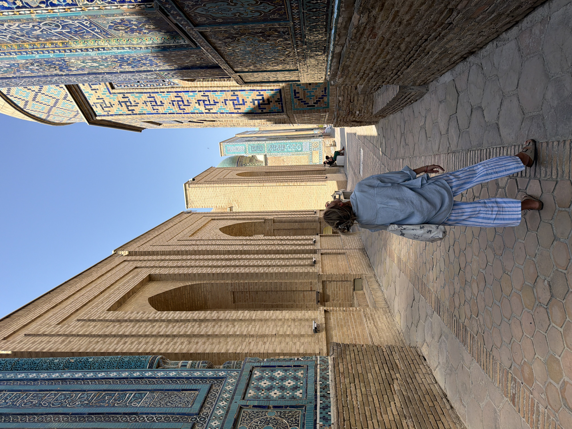 The image depicts a woman from the back, facing away from the camera, as she stands in a narrow alleyway in Samarkand, Uzbekistan. She is wearing a light blue, loose-fitting shirt and blue and white striped pants, with sandals on her feet. A tote bag is slung over her shoulder. Her hair is tied back in a bun. The alleyway is paved with hexagonal stone tiles and bordered by walls constructed of tan bricks. The architecture on either side of the alleyway features intricate blue tilework with Arabic inscriptions and geometric patterns, characteristic of historical Uzbek Islamic architecture. Arches are visible in the brick structures, and in the distance, a green dome is partially visible. Two other individuals are seated on a bench in the background, facing the woman. The sky is a clear, bright blue. The overall scene suggests a sunny day in a historically significant location.