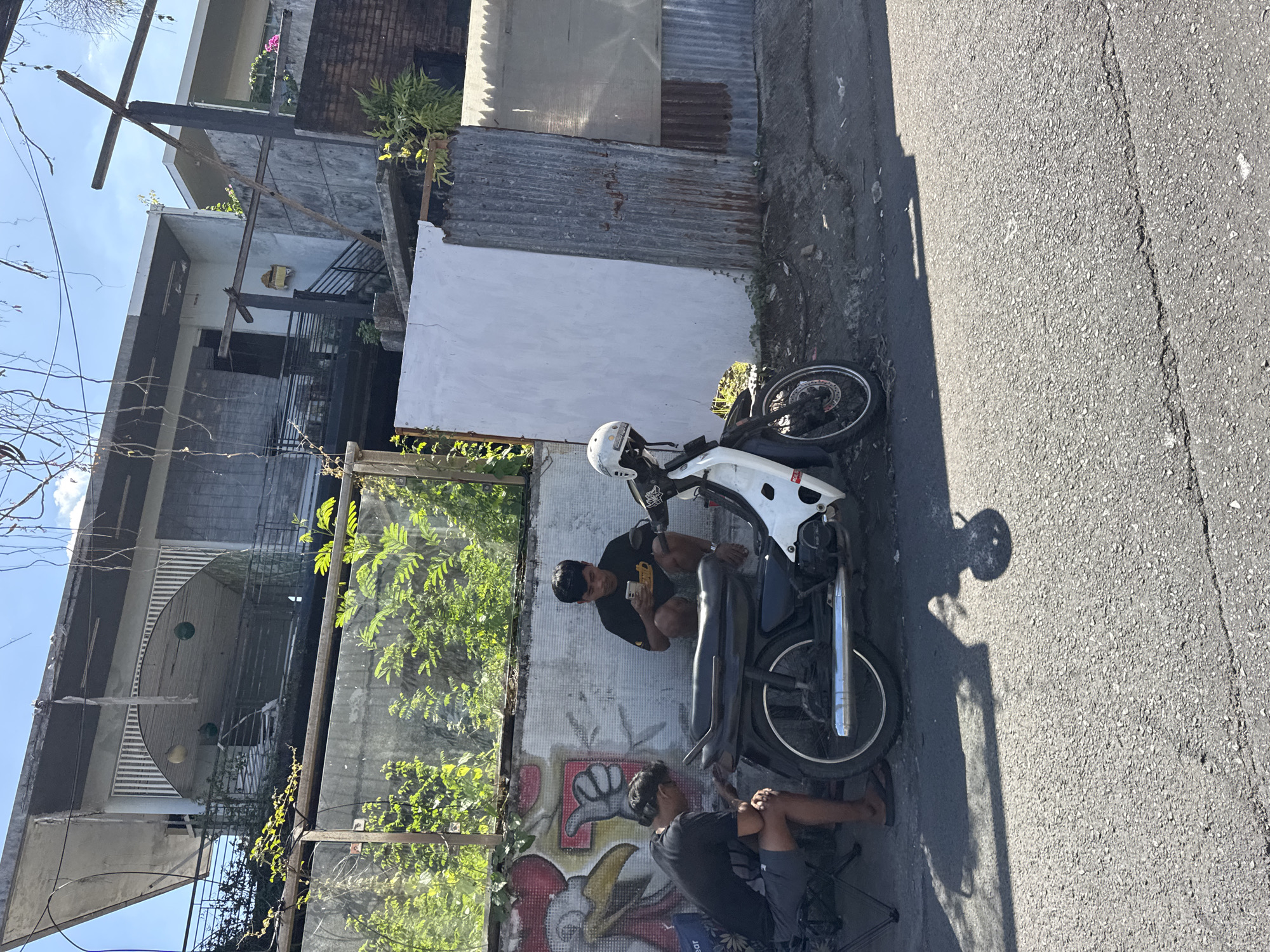 A medium shot captures a scene in Kuta, Indonesia. Two men are seated alongside a white and black motorcycle parked on the asphalt road. The first man is focused on his mobile phone. The second man is seated adjacent to the motorcycle with his knees drawn up. Both are seated against a graffiti-covered wall and vegetation. A building is visible in the background. The lighting suggests midday.