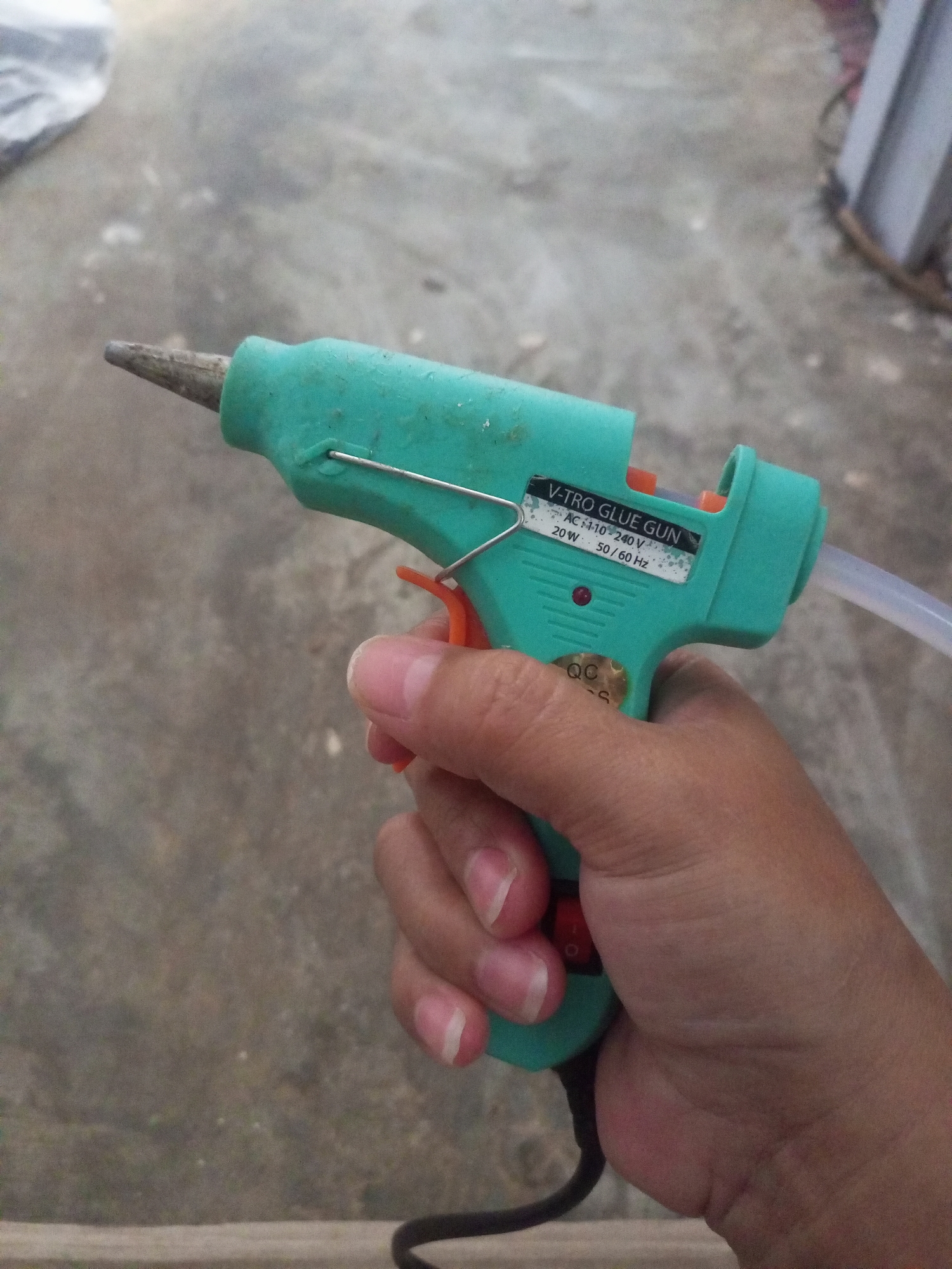 A person's hand is holding a green hot glue gun, with their index finger on the orange trigger. The glue gun has a label that reads "V-TRO GLUE GUN AC 110-240 V 20W 50/60 Hz". A clear tube extends from the back of the glue gun. The background is an outdoor setting with a concrete surface.