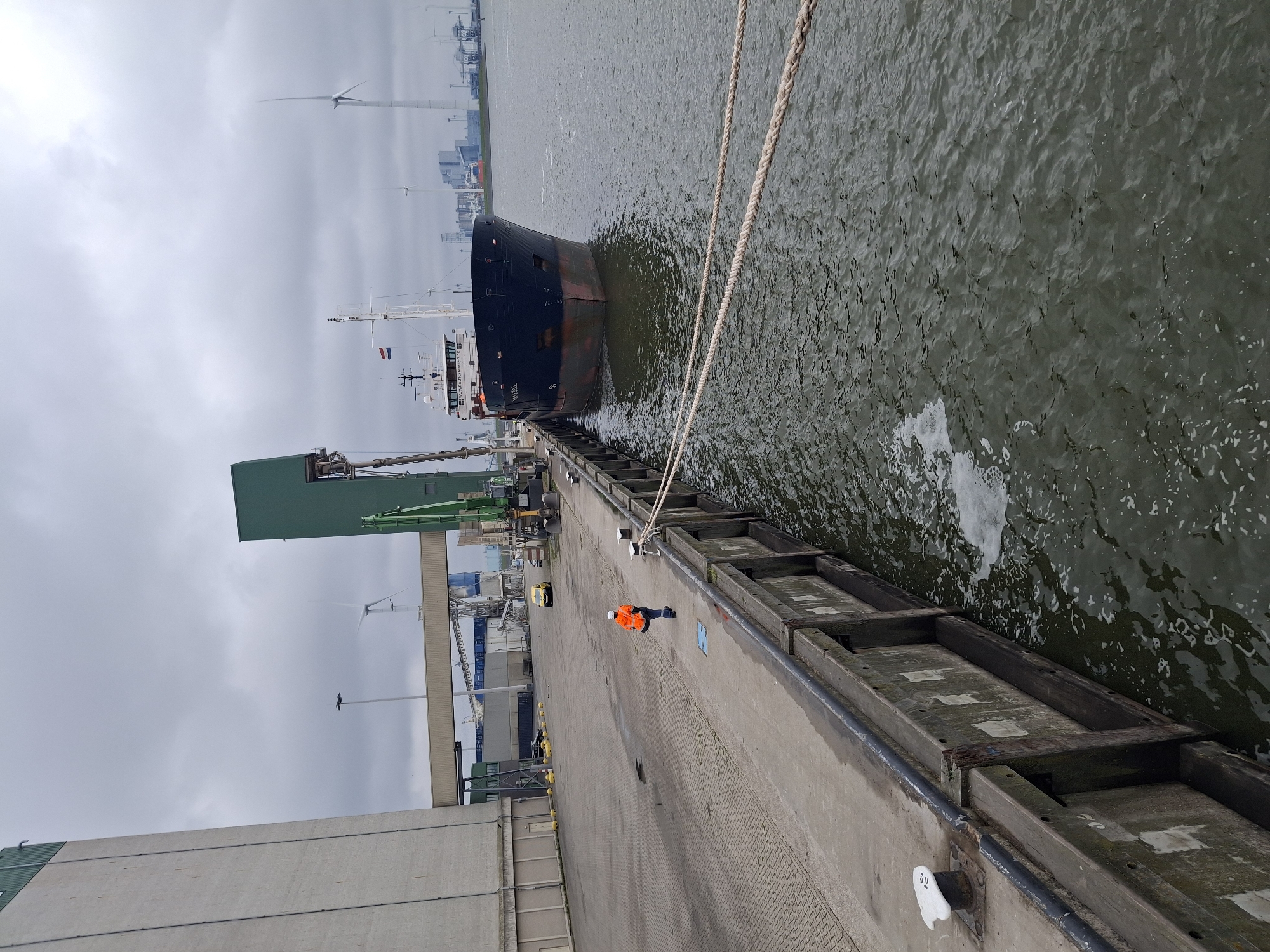 The image shows a cargo ship docked at the port in Eemshaven, Netherlands. A man wearing an orange safety vest is walking along the concrete pier beside the ship. The water is dark and choppy with some white foam. Ropes are connected between the ship and bollards on the pier. Cranes and industrial buildings are visible in the background, along with wind turbines in the distance. The sky is overcast.