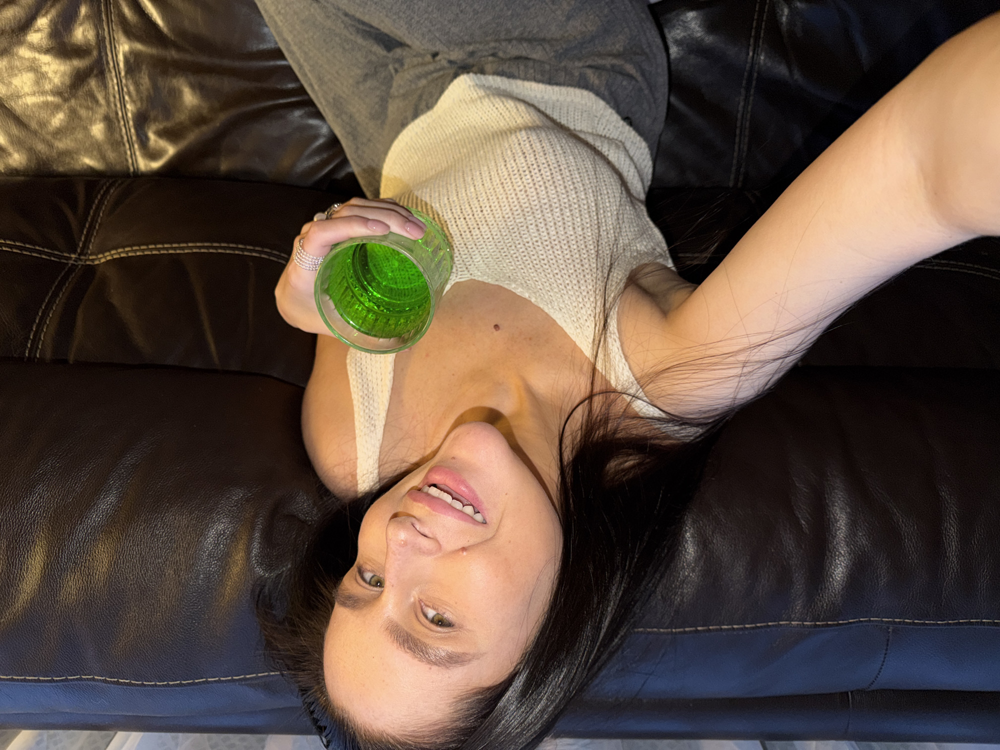 The image captures a young woman from an overhead perspective, reclining on a dark brown leather couch. She is looking upwards towards the camera, displaying a playful expression with a slight smile and a wink of her right eye. In her left hand, she holds a textured green glass containing a vibrant green liquid. She is dressed casually in a light-colored, possibly off-white, ribbed knit tank top and grey sweatpants. Her dark, long hair is spread out on the couch around her head. Rings are visible on the thumb and index finger of her left hand, which also features light pink manicured nails. The couch is dark brown leather with visible stitching. The scene is set indoors, likely a living room, under artificial lighting, suggesting an evening or nighttime context. There is no visible text in the image. The person appears relaxed and engaging. This scene is contextualized to Khimki, Russia.