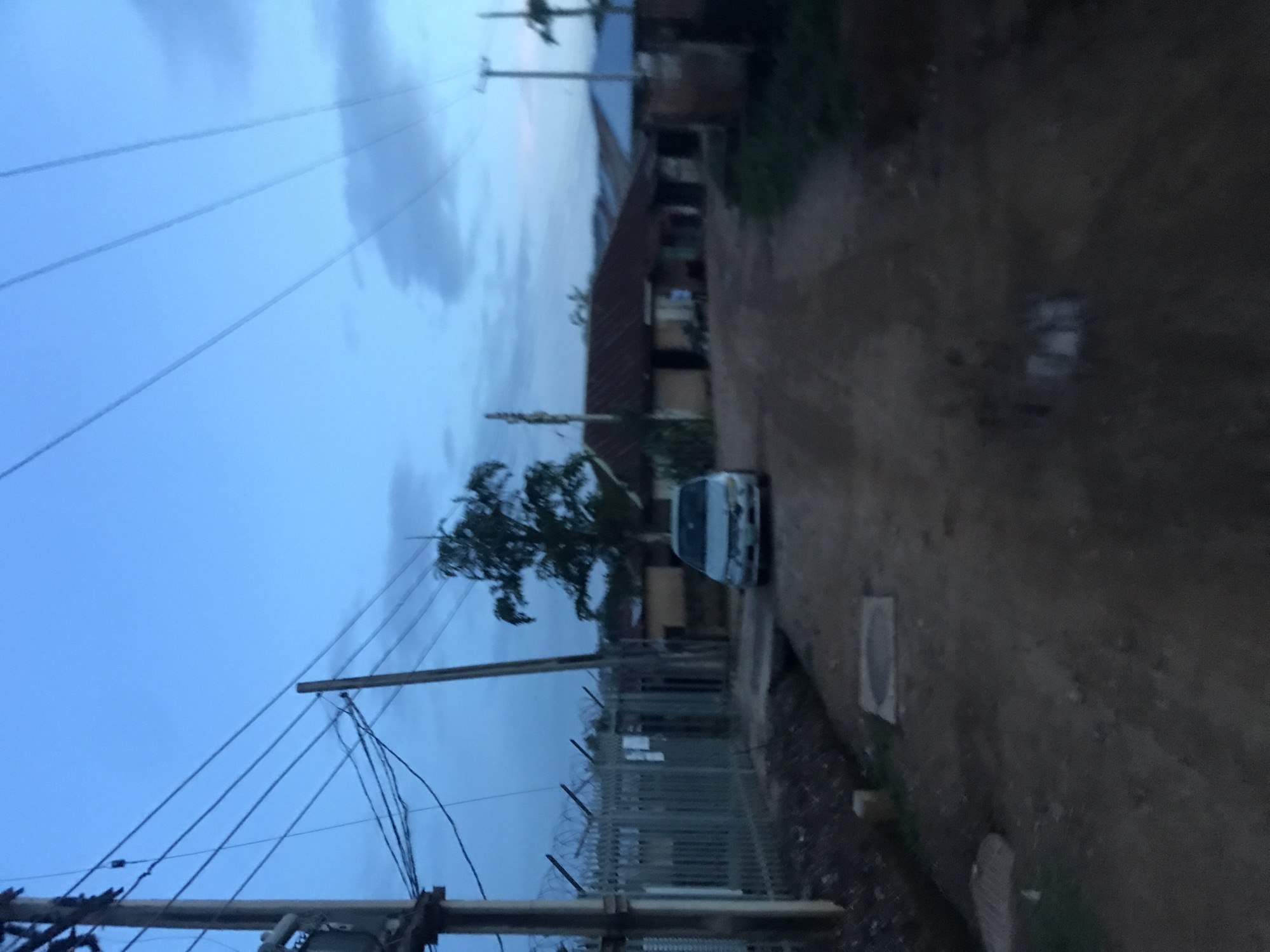 This image depicts a residential or semi-urban street in Rafin Par, Nigeria, observed during twilight hours, indicated by the subdued natural light and the blend of light blue and grey tones in the sky. The scene is dominated by an unpaved dirt road that recedes into the background.
In the foreground and mid-ground, a light-colored, possibly silver, sedan is parked centrally on the dirt road, facing the viewer. Its specific make and model are obscured by blur. To the left, a tall wooden utility pole stands prominently, supporting multiple overhead power lines that diagonally cross the upper portion of the frame. Adjacent to this pole, a metal fence topped with multiple strands of barbed wire encloses an area. Several smaller utility boxes or components are attached to the main utility pole. Further down the road, two rectangular utility covers are visible on the ground.
In the mid-ground, a large tree with dense, dark foliage stands to the right of the parked vehicle. Beyond the tree and the car, several single-story structures with distinct corrugated metal roofs are visible, indicating residential or commercial buildings. Another wooden utility pole is visible in the mid-ground on the right, also supporting power lines. The sky shows some scattered cloud formations. No clearly identifiable individuals are present in the scene.