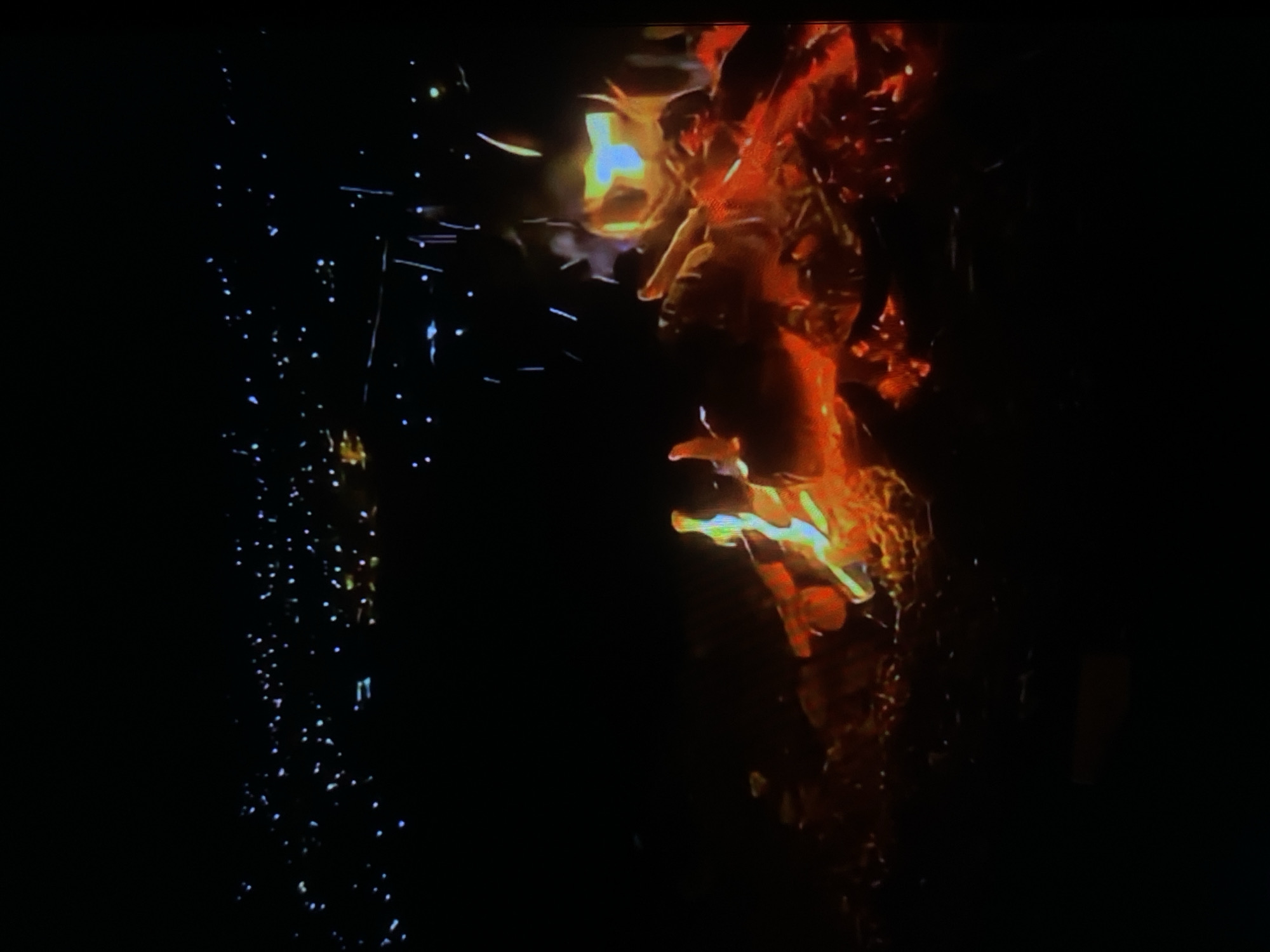 The image depicts a nighttime scene with a bonfire in the foreground and a distant city skyline. The bonfire is composed of logs and embers, with flames reaching upwards in shades of orange, yellow, and blue. Sparks are visible rising from the fire into the dark sky. In the background, a sprawling city lights up the horizon, with numerous small, bright dots suggesting buildings and streetlights. The city lights appear to extend across a vast area, indicating a significant urban or suburban landscape. The overall impression is one of a warm, inviting fire contrasting with the distant glow of a populated area under the night sky. There are no people or specific landmarks clearly identifiable in the image to pinpoint the exact location within Tinaquillo, Venezuela.