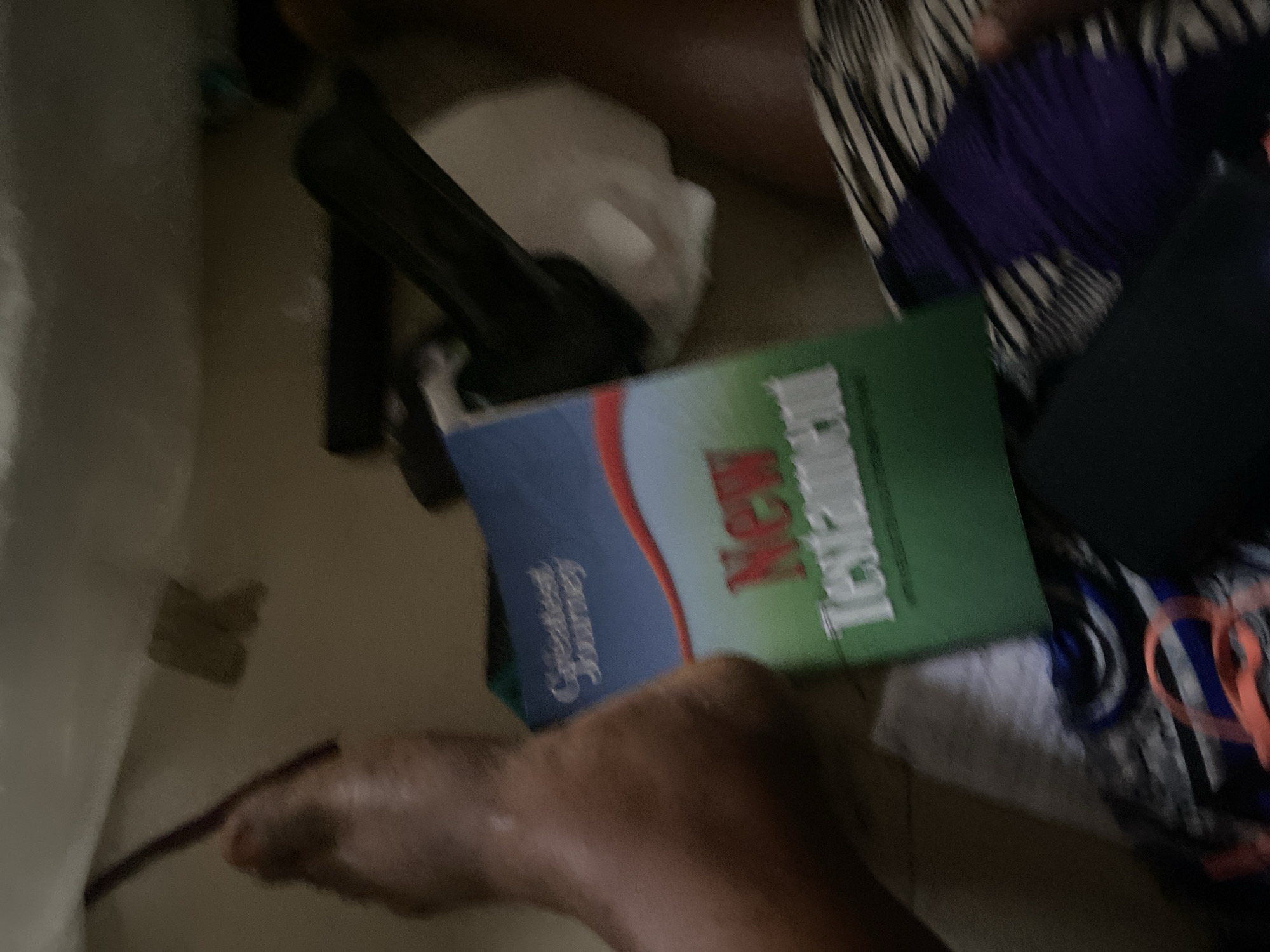 The image shows a close-up, slightly blurry view of a New Testament book with the title "Greatest Journey" above "New Testament" on its cover, lying on the floor amidst clothing and other objects. A person's foot and lower leg are visible on the left side of the frame, with a purple object, possibly a pencil or stylus, next to the foot. The background consists of a floor and what appears to be a bed or couch. The scene is cluttered, with a black object, possibly a remote or phone, and a crumpled white piece of paper also visible. The overall impression is of a casual, indoor setting. There is no information in the image to indicate its location within the city of Jauro Yinu, Nigeria.