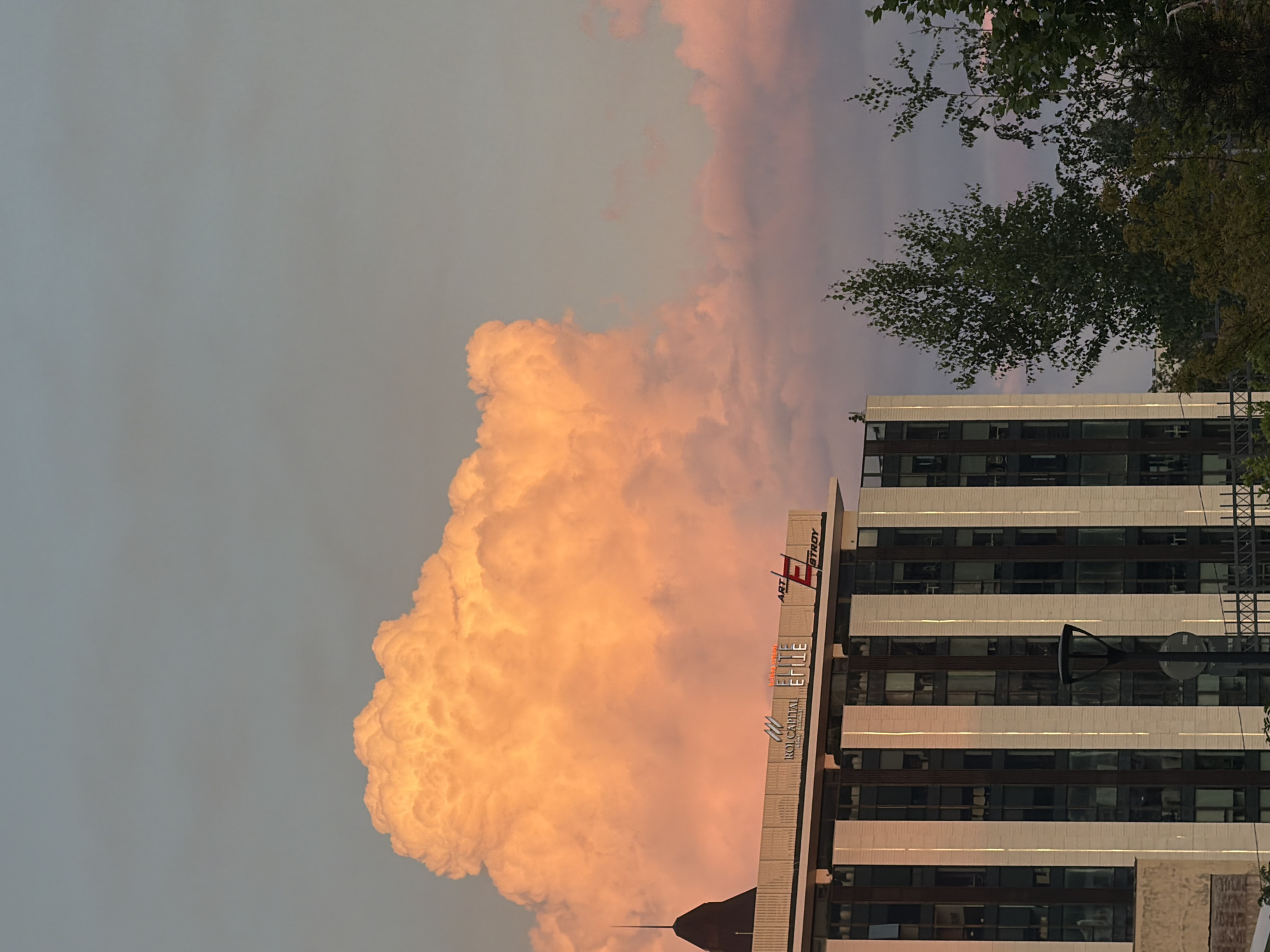 The image captures a building in Sofia, Bulgaria, against a dramatic sky filled with a large, luminous orange cloud. The building, which appears to be an office or commercial structure, features a modern facade with vertical white elements and numerous dark-tinted windows. The top of the building displays signage, including "ROI CAPITAL," "ELITE," and "ART ESTROY," indicating businesses located there. A dark, pointed spire-like structure protrudes from the left side of the building's roof. In the foreground and to the right, several trees with green foliage partially obscure the lower portion of the building and extend upwards. The sky transitions from a soft blue-gray at the top to hues of pink and orange, particularly around the prominent cloud, suggesting either sunrise or sunset. A street lamp is visible in front of the building.