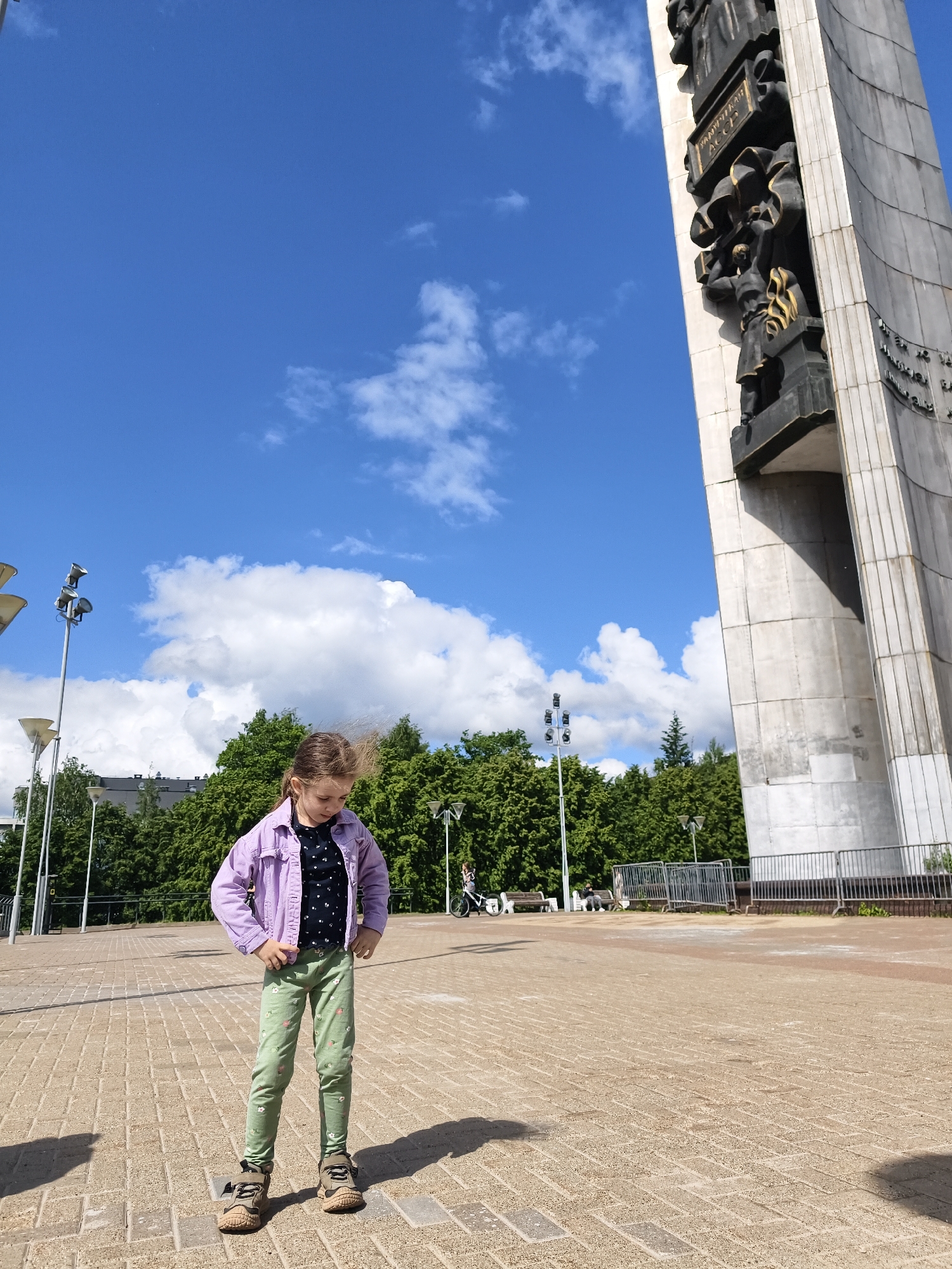 A young girl stands prominently in the foreground of a large outdoor paved square, casting a shadow to her right. She is wearing a light purple denim jacket, a dark blue t-shirt with light polka dots, and light green patterned leggings, with brown athletic shoes. Her hair, light brown, appears to be slightly windblown. She is looking downwards, with her hands resting on her hips.
To the right, a massive concrete monument rises vertically, featuring intricate bronze relief carvings depicting figures and historical scenes. One visible plaque on the monument clearly displays the Cyrillic text "УДМУРТСКАЯ АССР" (UDMURTSKAYA ASSR), suggesting a historical or commemorative significance related to the Udmurt Autonomous Soviet Socialist Republic.
The background features lush green trees, streetlights, and some distant buildings. Further back on the right side of the square, fences and what appears to be other people or benches can be seen, along with a bicycle. The sky is bright blue with scattered white, fluffy clouds, indicating clear and sunny weather during the daytime. The overall scene suggests a public square or park area in Izhevsk, Russia, likely a historical or memorial site given the monument. No specific activity is taking place beyond the girl standing in the square and other distant figures.