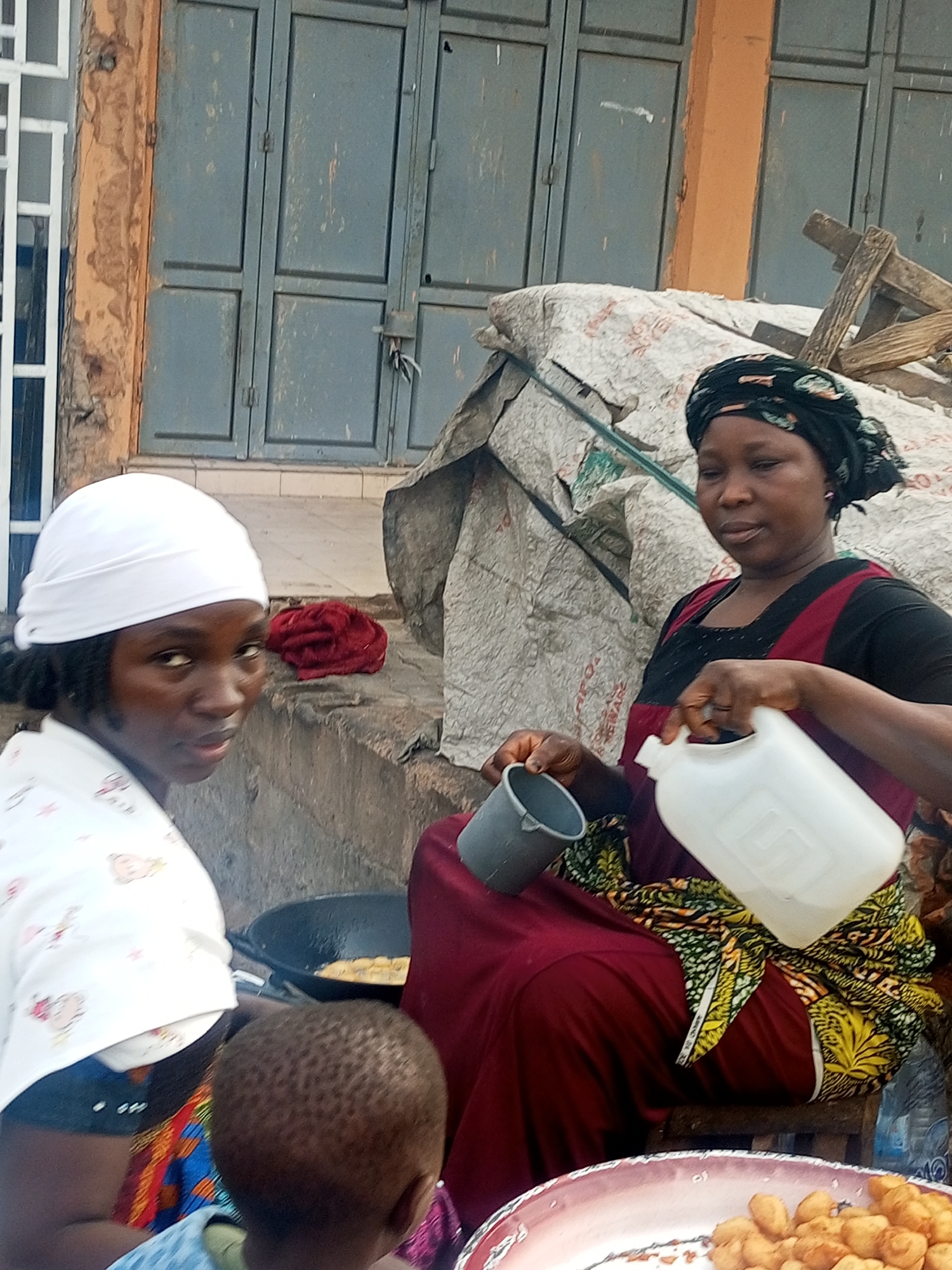 In Tempure, Nigeria, two women are shown preparing food outdoors. One woman, wearing a white headscarf and a light-colored, patterned top, looks towards the camera. The second woman, with a black head covering, sits on a stool and pours liquid from a white plastic container into a gray cup. She is wearing a maroon dress and a colorful patterned skirt. A young child is partially visible in the foreground. A black pan with frying food is visible behind the woman in the white headscarf. A tray of fried dough balls is in the foreground. Behind the women, there's a wall with closed, grayish-blue doors and a tarpaulin-covered structure.