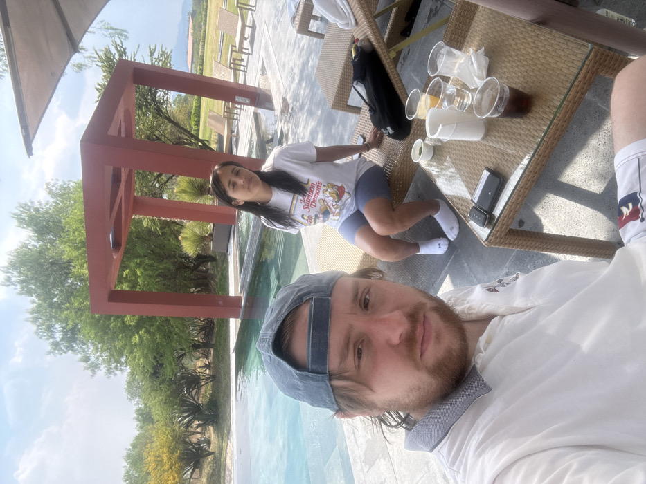 This outdoor image, captured in Pueblo Nuevo de Jasso, Mexico, depicts two individuals beside a swimming pool under a clear, sunny sky.
In the foreground, a light-skinned male with a beard and a blue baseball cap worn backward is visible from the chest up, taking a selfie. He wears a light-colored polo shirt with a contrasting collar. His lower body is partially visible, showing patterned shorts.
To the right, in the mid-ground, a female with dark, straight hair is seated on a light-brown wicker lounge chair. She wears a white t-shirt with a graphic print, blue shorts, and white ankle socks. Her posture is relaxed, with her head turned slightly towards the camera.
Between the two individuals, a low, rectangular wicker table with a glass top holds several items. These include multiple plastic cups, some containing beverages (one dark, one orange, one clear), a small green container, a small white container, a dark-colored smartphone or electronic device, and a crumpled white napkin.
Behind the female, another wicker lounge chair is visible, supporting a black bag. The swimming pool, featuring blue water and dark submerged steps, extends behind the individuals. Beyond the pool, a prominent, open-frame, rust-red architectural structure stands. The surrounding area is paved with light-colored tiles.
The background consists of lush green trees and shrubs, including some with yellow blossoms. In the far distance, low hills or mountains are visible under a partly cloudy sky, along with a building featuring a reddish roof on the right. A portion of a large, light-colored overhead canopy is visible in the upper right corner.