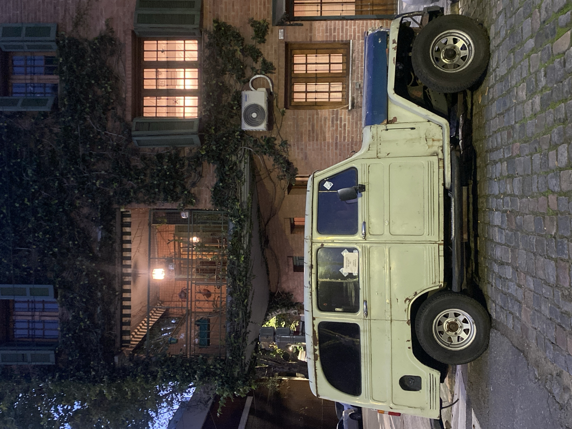 An exterior shot reveals a vintage vehicle parked along a cobbled street beside a brick building, likely in a historical district. The building is adorned with ivy, and windows are lit. An air conditioning unit is affixed to the wall. No people are immediately visible. It's strongly suggested that this is in Buenos Aires, Argentina, because of the architectural style of the brick building and the old-fashioned vehicle, both very common in that area. The image is taken at dusk or night.