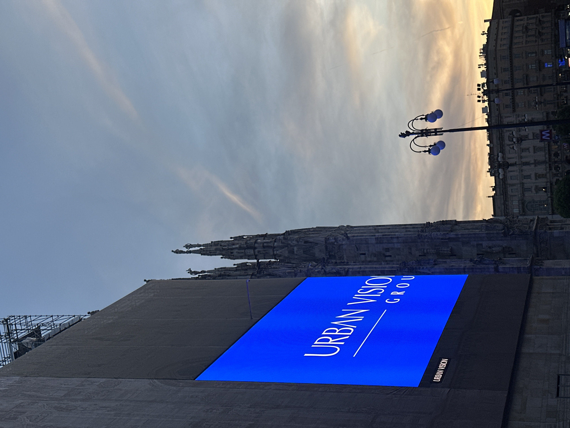 This is a medium-shot, eye-level photograph taken at dusk in Milan, Italy, specifically in front of the Duomo. The main subject is a large, brightly lit blue LED screen displaying white text that reads "URBAN VISION GROUP". Below this, a smaller version of the logo, "URBAN VISION", is visible. To the right of the screen, the intricate Gothic spires of the Duomo are silhouetted against the twilight sky, which is a gradient of pale blue and orange hues with wispy clouds. In the foreground, a vintage-style streetlamp with multiple globes, some illuminated with a blue light, stands as another prominent object. Further in the background, the facades of classical European buildings are visible, with some lights on. The scene is relatively devoid of people, with only a few indistinct figures visible on scaffolding atop the building to the left and on the Duomo's spires. The overall atmosphere is serene and urban, capturing a moment of transition between day and night in a historic city square.