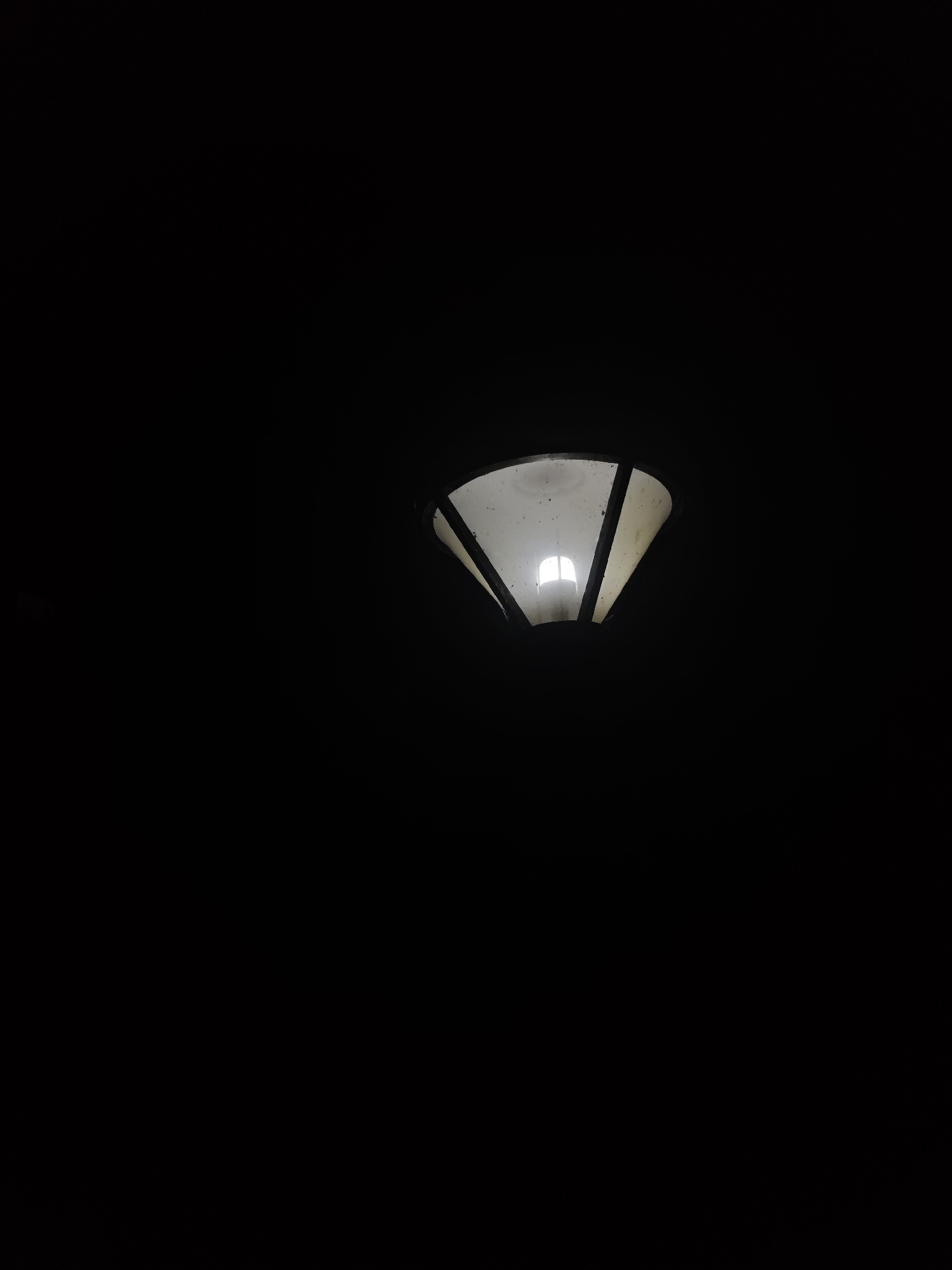 The image shows a close-up, low-angle shot of a street lamp at night. The lamp is hexagonal in shape with white translucent panels and black dividers. A single light bulb illuminates the inside of the lamp, casting a soft glow through the panels. The background is entirely black, indicating a very dark environment. The scene is devoid of any people or other discernible objects. No specific location in the Chongqing High-tech Zone, China, can be identified from this image.