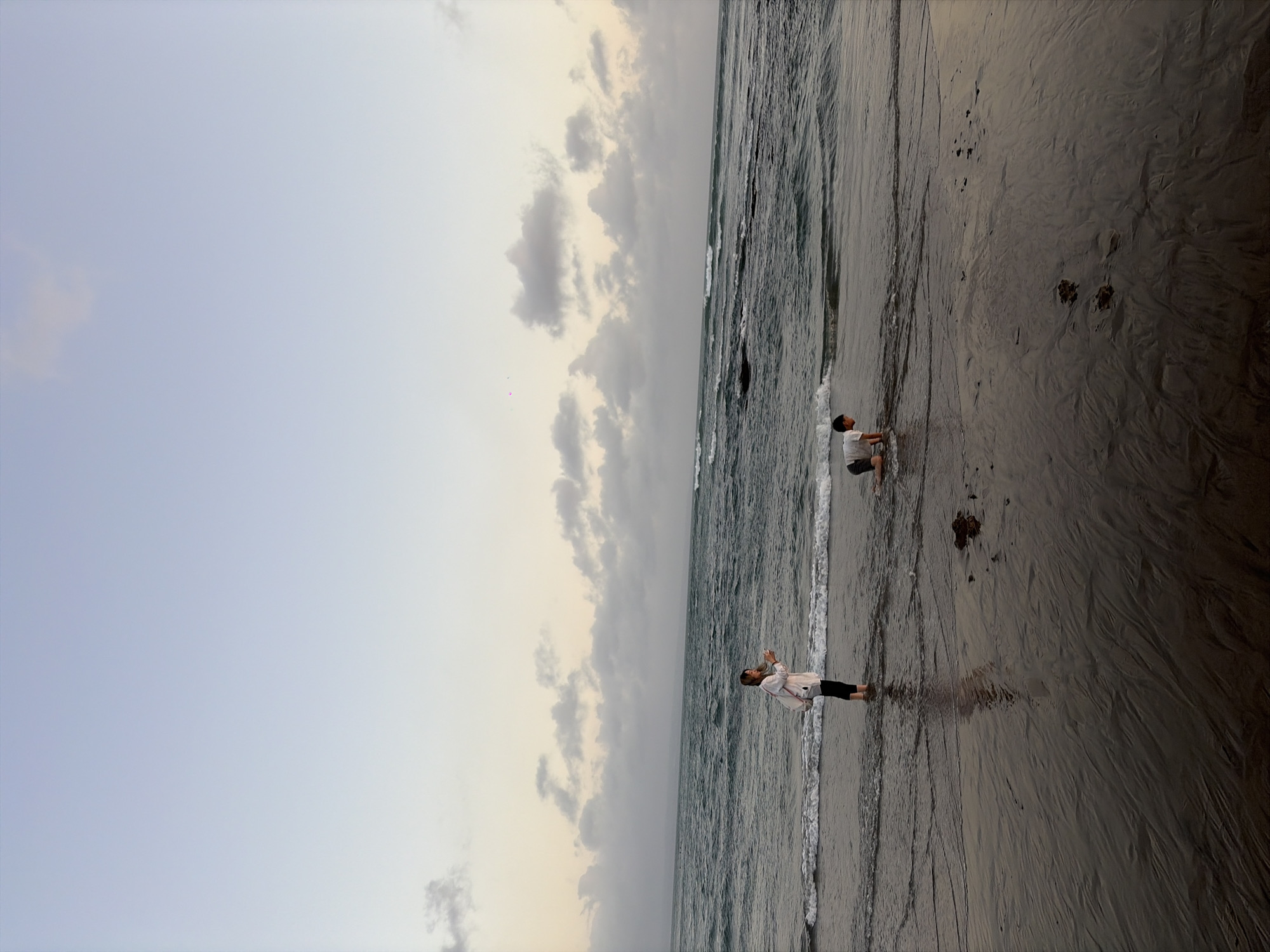 At dusk on a beach in North Kuta, Indonesia, a woman stands knee-deep in the ocean, holding a white cup and a bag. To her right, a young boy kneels in the shallow water, facing the sea. The tide is out, exposing wet sand with ripples and some debris. The sky is a pale gradient of blue and yellow, with scattered clouds. Gentle waves roll towards the shore, and the ocean appears dark and choppy. The overall scene suggests a peaceful evening by the sea.