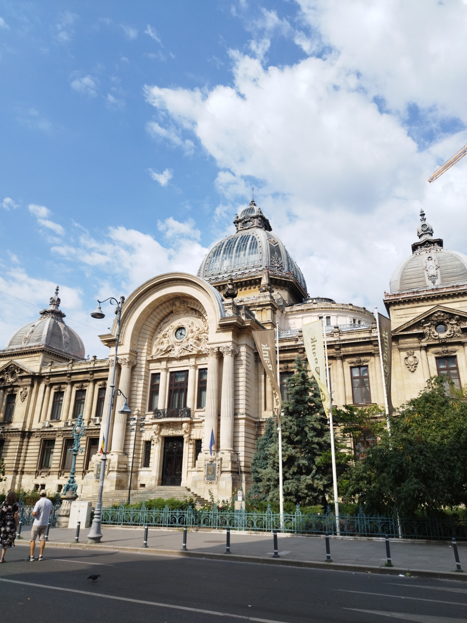 The image shows the National Bank of Romania building in Bucharest, Romania. The grand Beaux-Arts building features multiple domes, ornate stonework, and large arched windows. In the foreground, a street with a pedestrian sidewalk separates the viewer from the building. Two people are walking on the sidewalk, and a bird is on the street. A decorative fence lines the edge of the building's property, and several tall evergreen trees are planted behind it. The sky above is blue with scattered white clouds, and a construction crane is visible in the distance.
