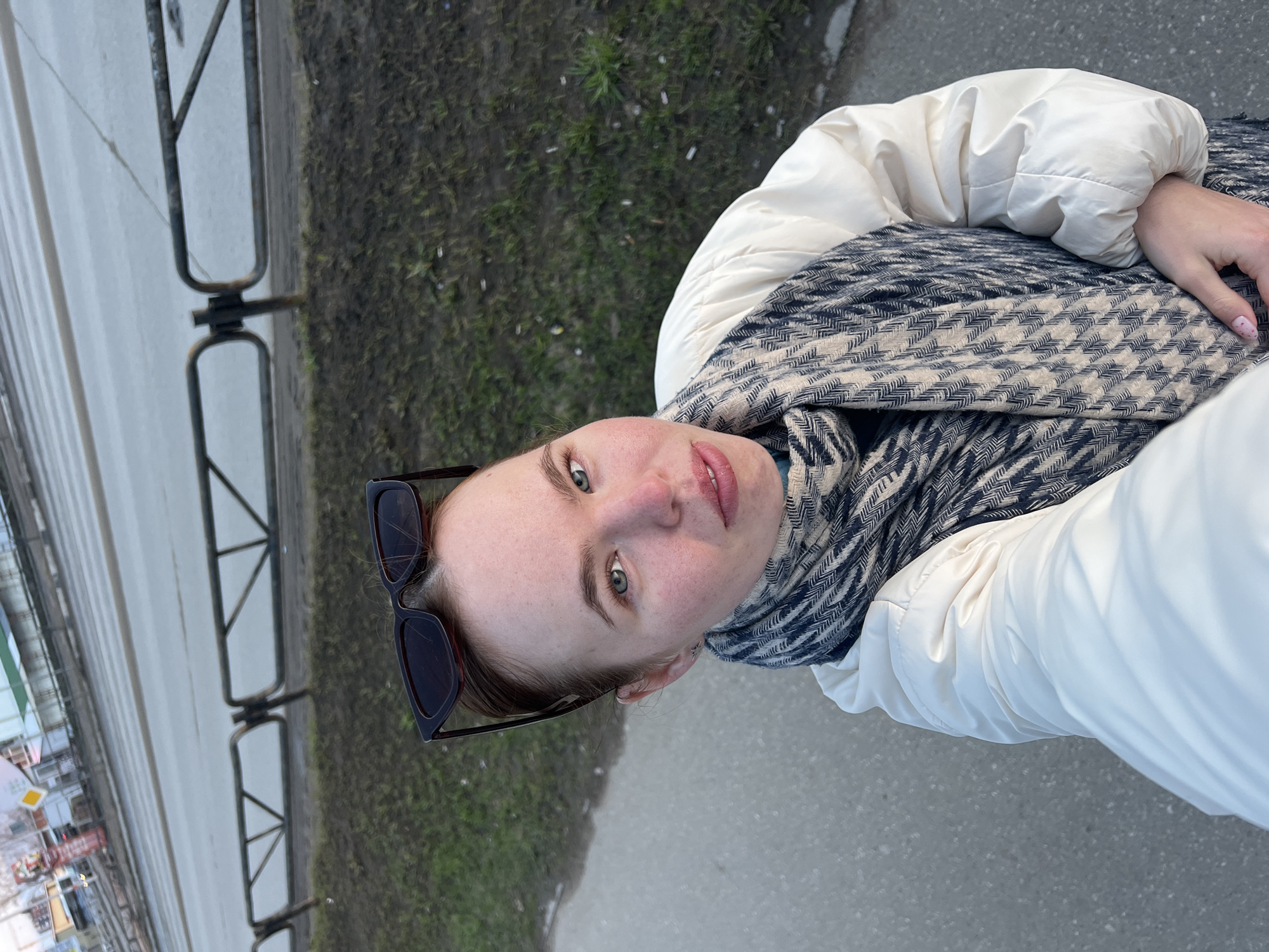 A woman stands outdoors on a cloudy day. She wears a white puffer jacket and a patterned scarf. Sunglasses rest on her forehead. Behind her is a gray concrete path, a patch of green grass, and a metal barrier fence. Across the street, blurry structures and signs suggest a cityscape. Based on the presence of Russian signage and architecture, this scene is plausibly located in Chaikovsky, Russia.