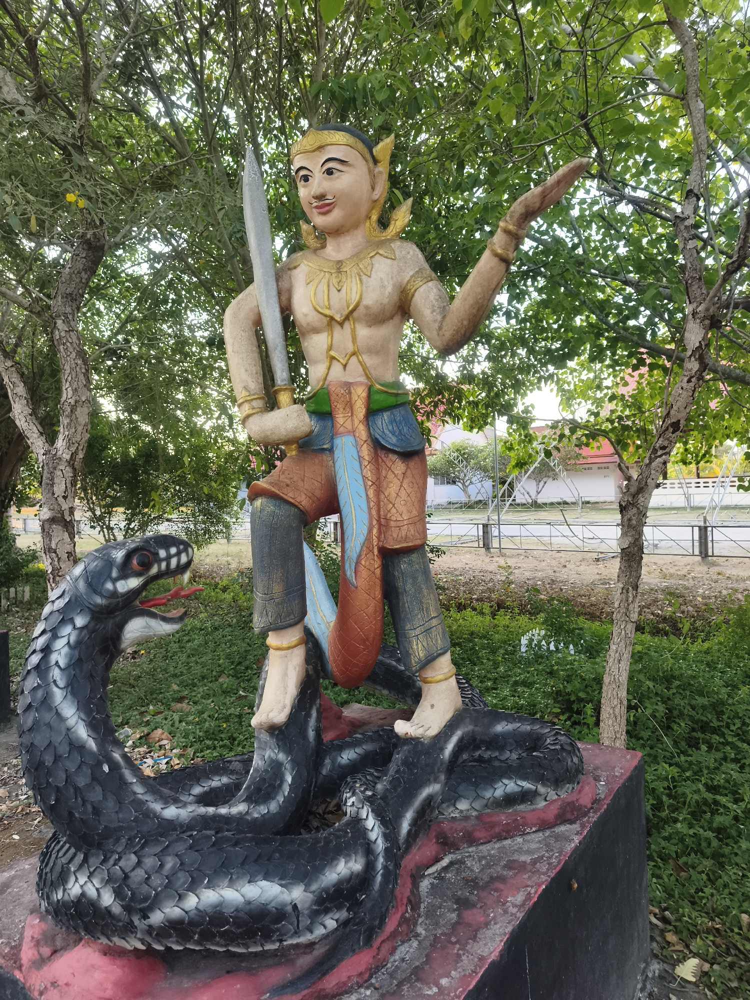 The image depicts a colorfully painted concrete statue of a male figure with a sword, standing atop a coiled snake. The figure has brown skin, gold ornamentation around the chest and head, and is wearing patterned pants. The snake is predominantly black with a red mouth. The statue is located outdoors amidst greenery and trees, which are common to the urban landscaping of Baan Bo Phut, Thailand. A fence and buildings are visible in the background, further suggesting an urban or suburban setting in Baan Bo Phut.