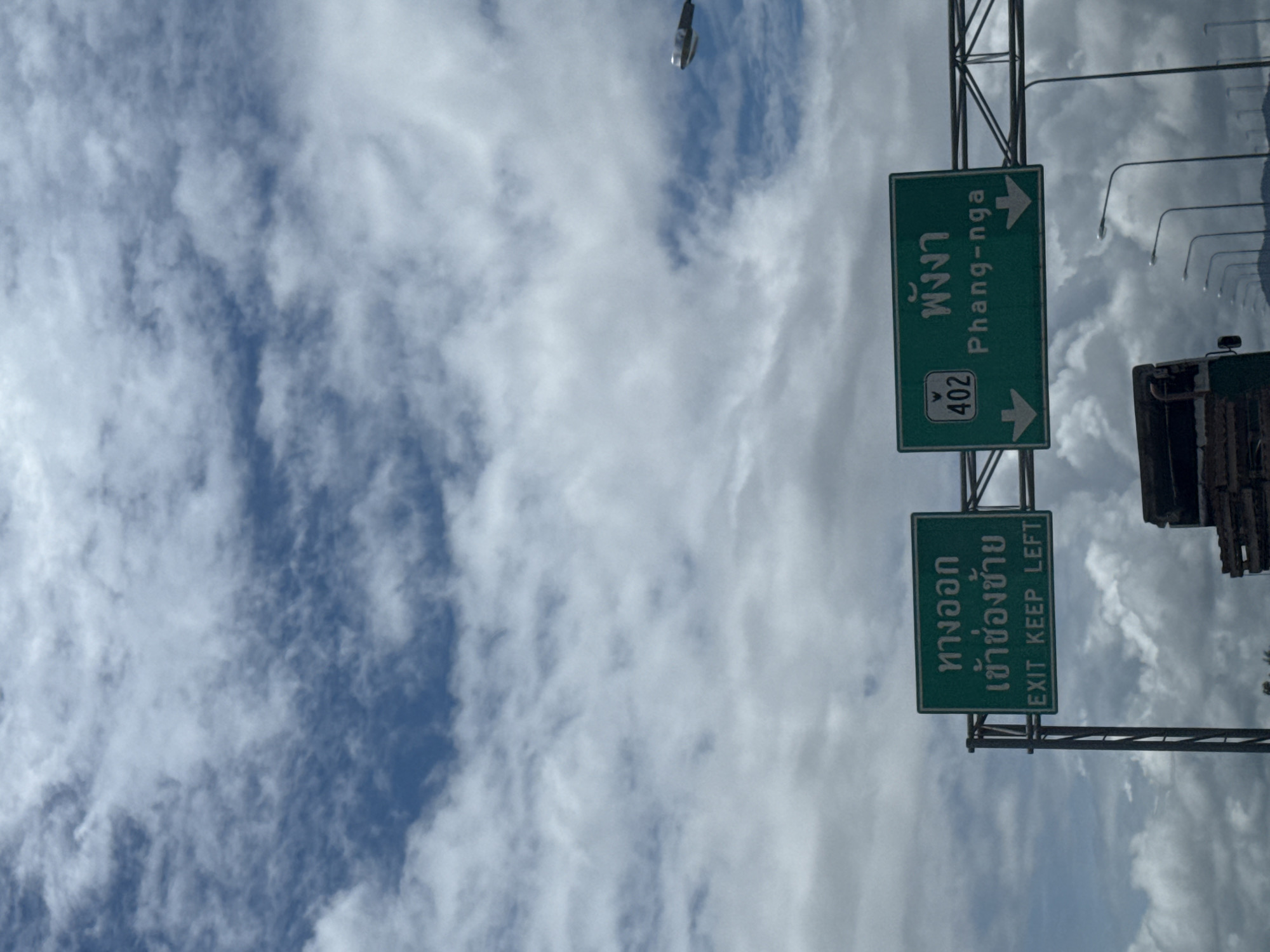 A highway signpost stands against a backdrop of a cloudy sky. The signpost has two green signs. The top sign, indicating "Phang-nga" and highway number 402, has an arrow pointing to the right. The bottom sign, in Thai and English, reads "EXIT KEEP LEFT" with an arrow pointing to the left. The clouds are white and fluffy, suggesting a daytime scene. The context provided indicates the location is Ban Tha Nun, Thailand. There are no people visible in the image.