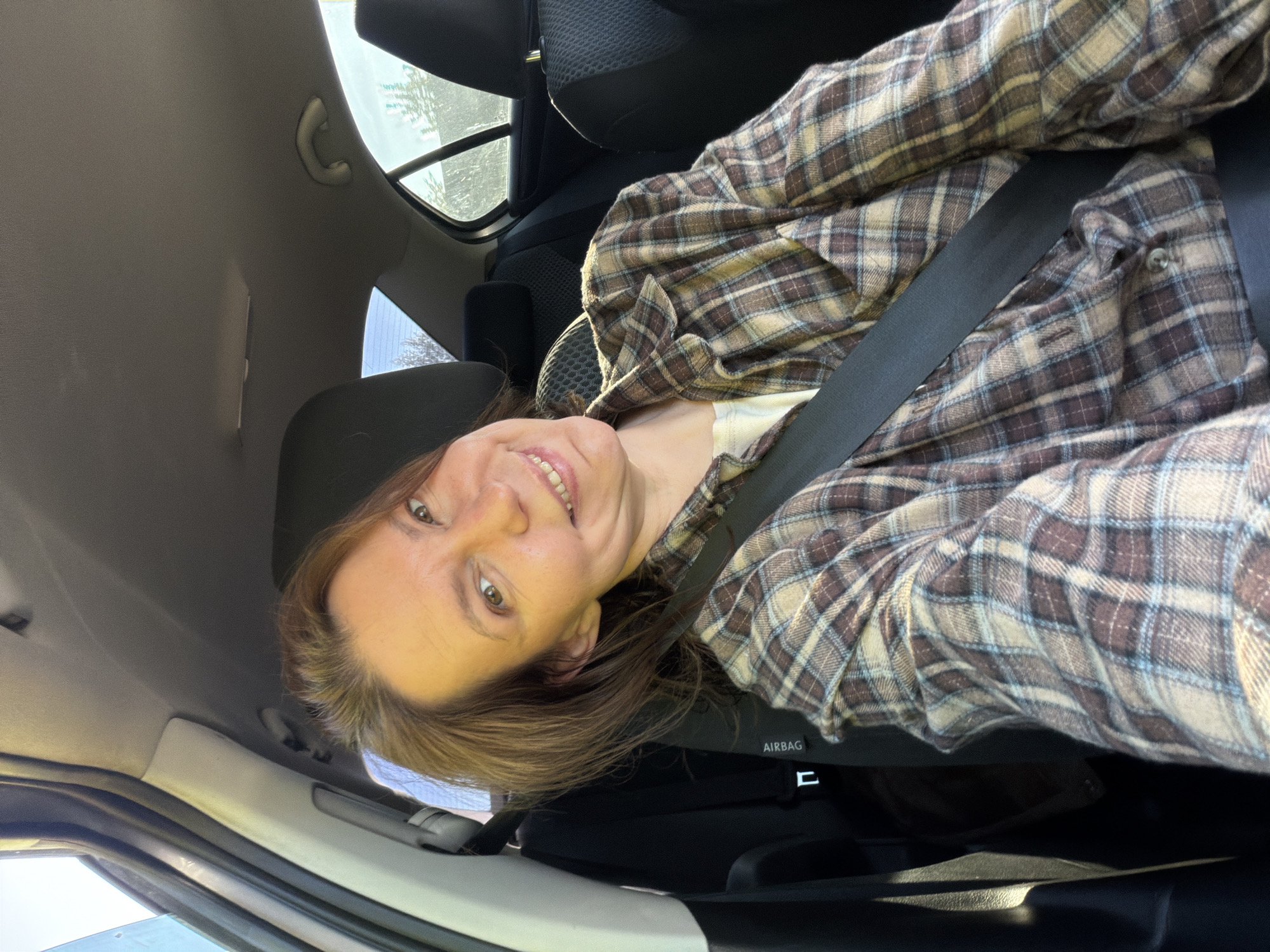 A rotated, eye-level shot shows a woman seated in the passenger side of a car, smiling warmly at the camera. The image is taken from a slightly low angle, looking upwards towards the woman's face and the car's interior ceiling.
The woman has light brown hair and is wearing a brown, beige, and light blue plaid flannel shirt over a white undershirt. A black seatbelt crosses her chest. Her face is well-lit, and she has a noticeable smile, revealing her teeth.
The setting is the interior of a car. Elements of the car visible include the dark fabric seats, the dashboard, the headliner, and a grab handle on the ceiling. Through the car windows, blurred glimpses of the outside environment can be seen, suggesting trees or foliage and bright daylight. The strong light indicates a sunny day. A distinct label "AIRBAG" is visible on the passenger side dashboard. The overall impression is one of a casual moment captured inside a vehicle during the day.