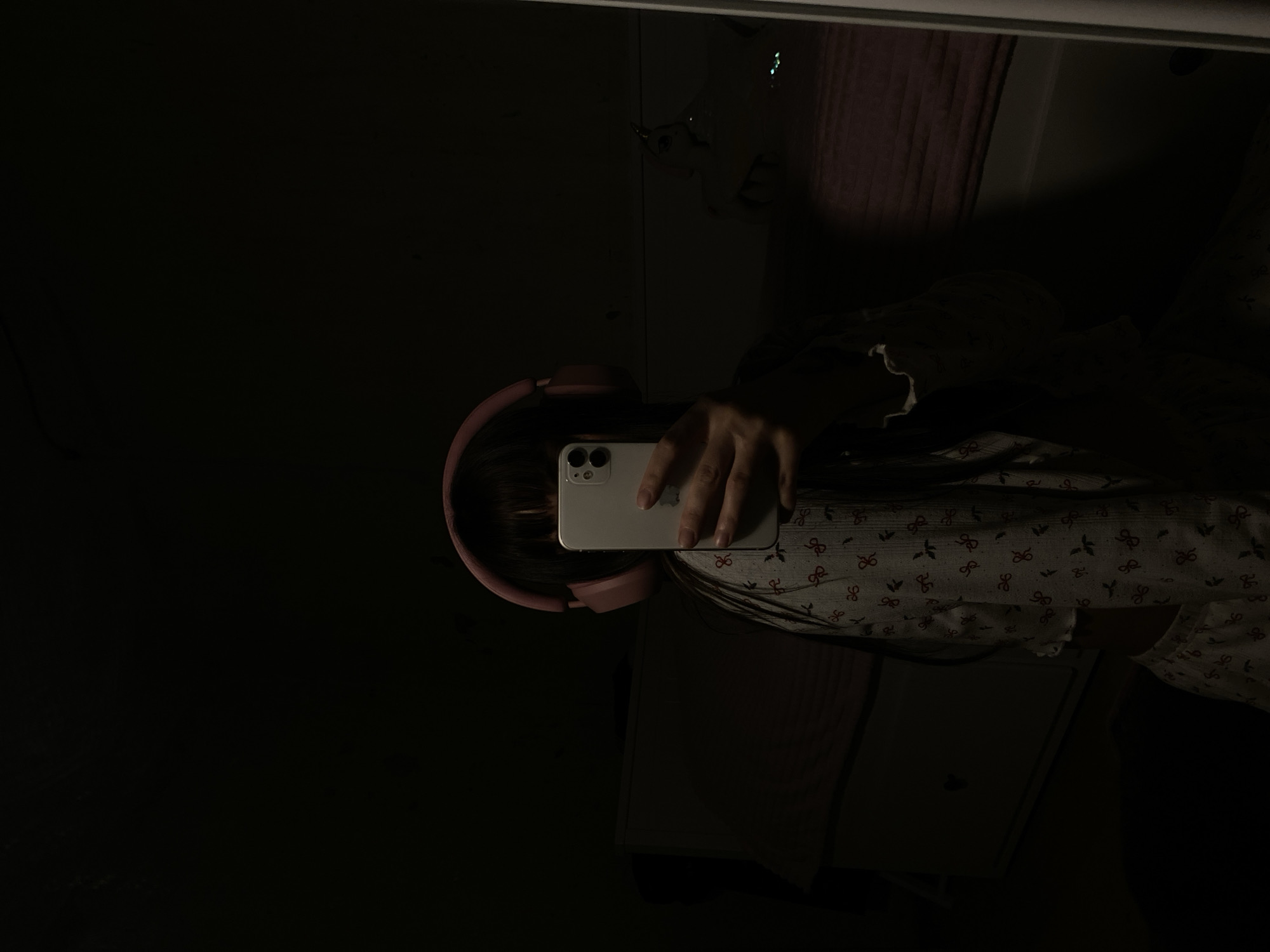 A person is taking a mirror selfie. The person is wearing pink headphones. They are holding a white phone in front of their face. The person is wearing pajamas with a small bow pattern. A dark purple curtain is partially visible behind the person in the mirror. The scene is dark and the location cannot be determined from the image.