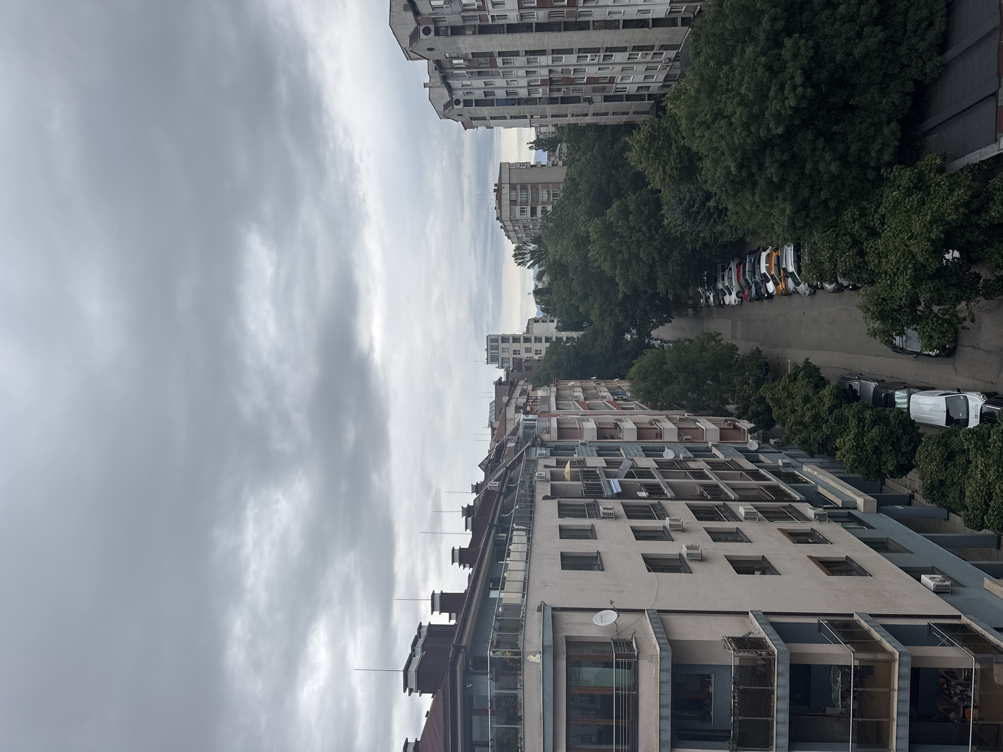 An aerial perspective captures a cloudy day over a residential street in Sofia, Bulgaria. The view looks down a street lined with apartment buildings on both sides, interspersed with mature green trees. The buildings exhibit a mix of architectural styles, with some featuring modern balconies and others more traditional facades. Parked cars are visible along the street and in designated parking areas. The sky is overcast, contributing to a muted color palette across the scene.