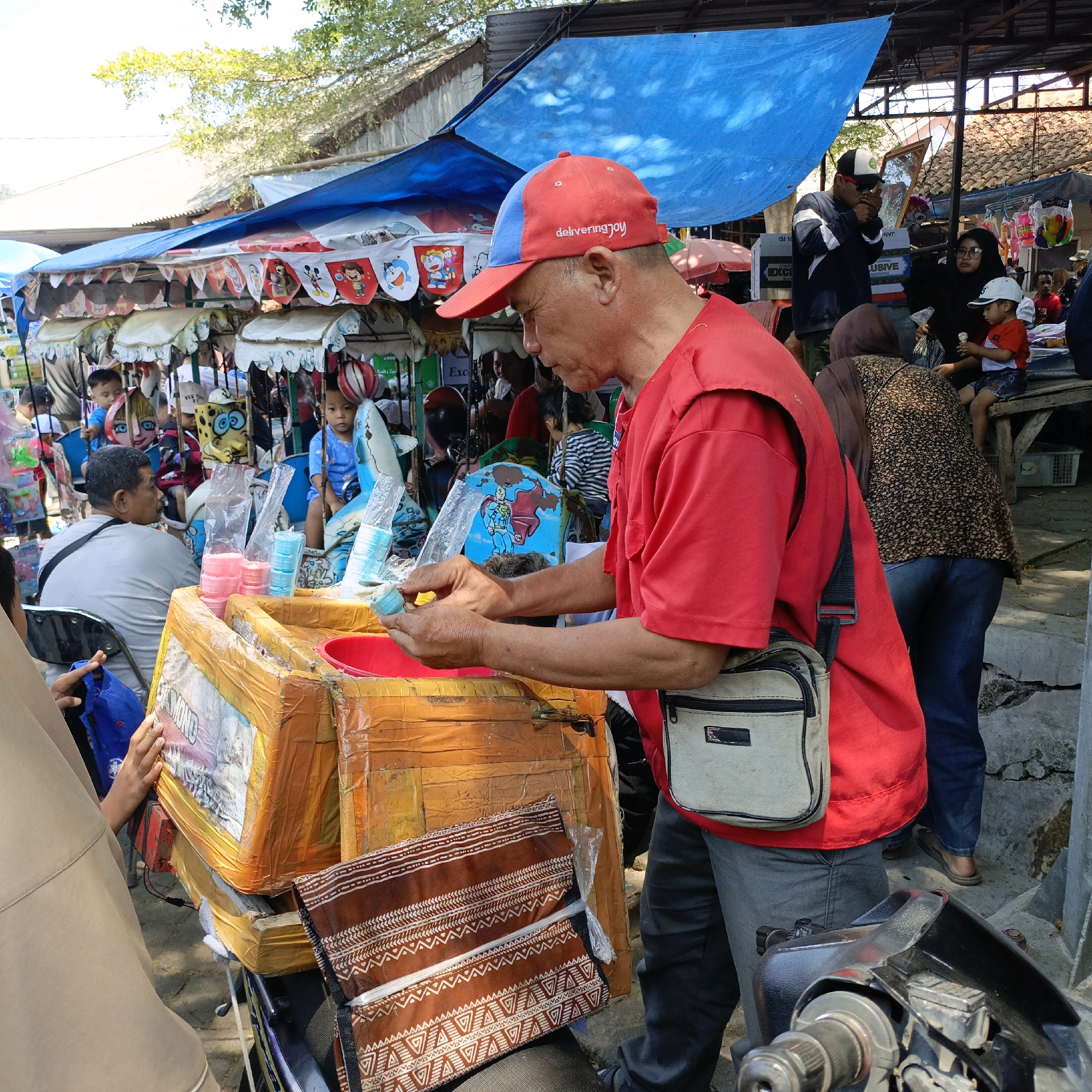 A man in a red shirt and a red and blue baseball cap is preparing ice cream for children at a market in Ambarawa, Indonesia. He is holding a blue ice cream cone and appears to be in the process of adding topping or a wrapper. In the background, children are gathered around various market stalls selling toys and treats. A blue tarp covers part of the market, and trees are visible overhead. The scene depicts a lively street market atmosphere with a focus on the ice cream vendor and his young customers.