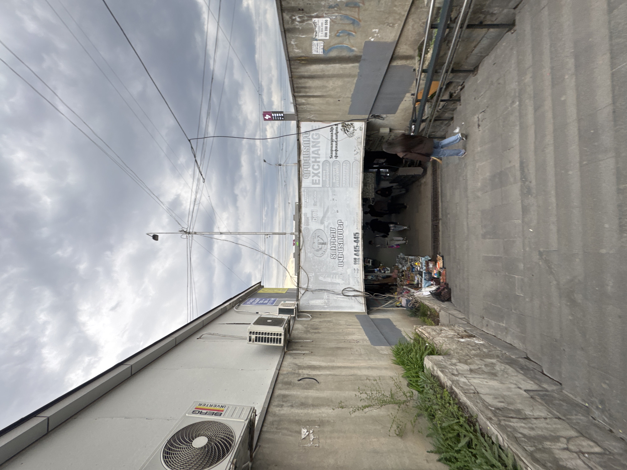 The image presents a view of a street scene in Yerevan, Armenia, under a cloudy sky. In the foreground, the side of a building is visible, featuring an air conditioning unit with the brand name "BERG INVERTER". Above it, two additional air conditioning units are mounted on the wall. A large sign is displayed, indicating a "MONEY EXCHANGE" and an "Aviatickets". This establishment provides currency exchange services. A person is walking on the sidewalk. Next to the sidewalk are some steps, with vegetation growing along the edges. Street cables stretch overhead.