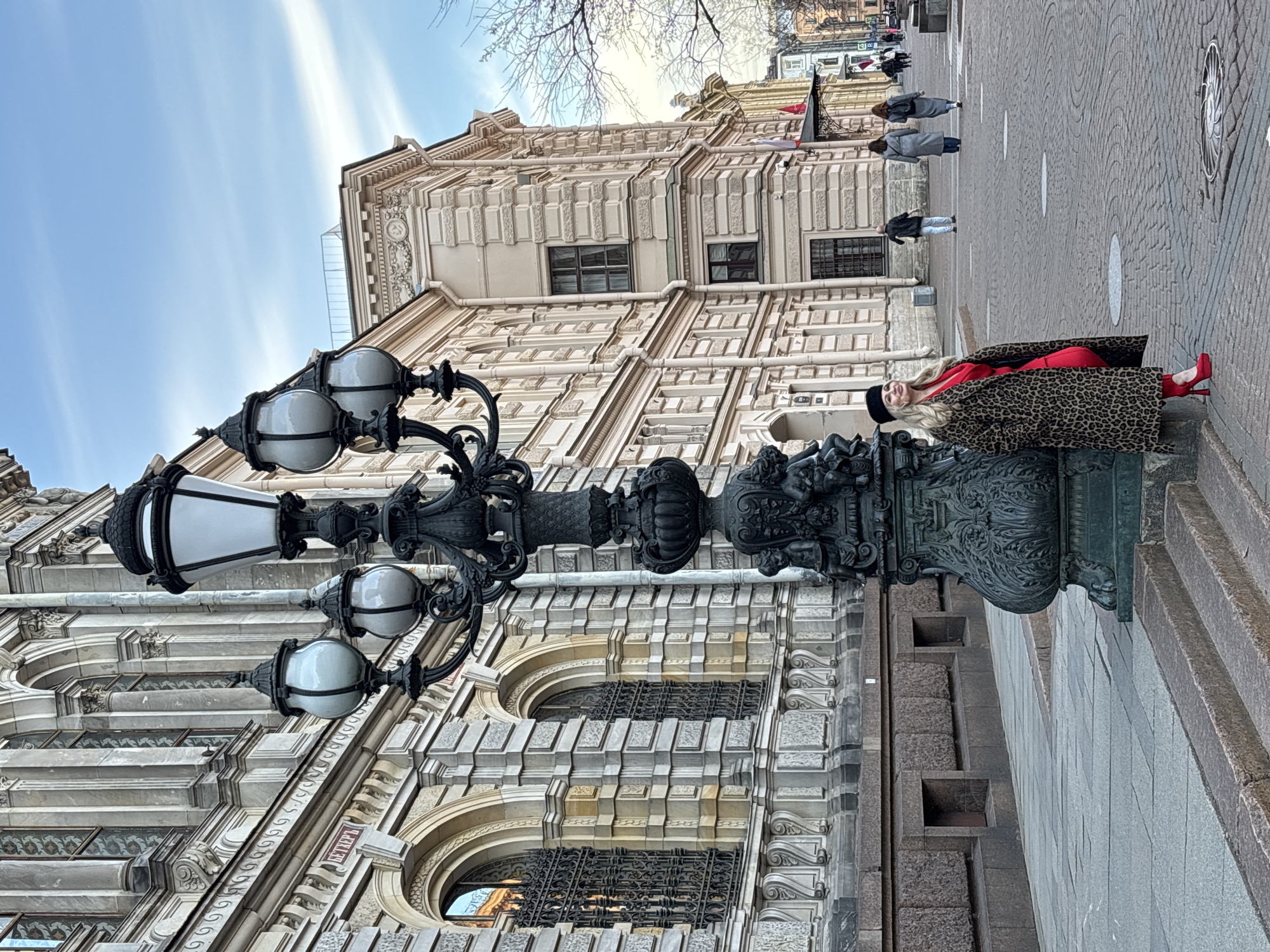This is an outdoor shot taken in Saint Petersburg, Russia. In the foreground is an ornate black lamppost with multiple lights. A woman is reclining against the base of the lamppost. She is wearing a leopard-print coat over a red outfit and red high heels. She is also wearing a black hat. In the background, there is a large, elaborate building with intricate architectural details. Several pedestrians are walking on the sidewalk in the distance. The sky is blue with streaky clouds.