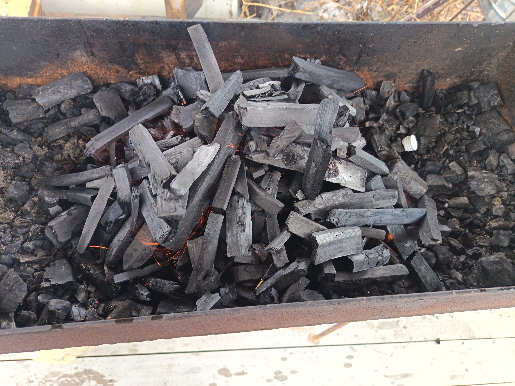 The image shows a metal box filled with charcoal and wood kindling, some of which are actively burning with visible sparks. The box appears to be a barbeque grill or fire pit. The background is blurry but seems to be an outdoor environment. There is a white cylindrical object on the right side of the image. The scene is likely related to preparing a fire for cooking or warmth. The location is unknown.