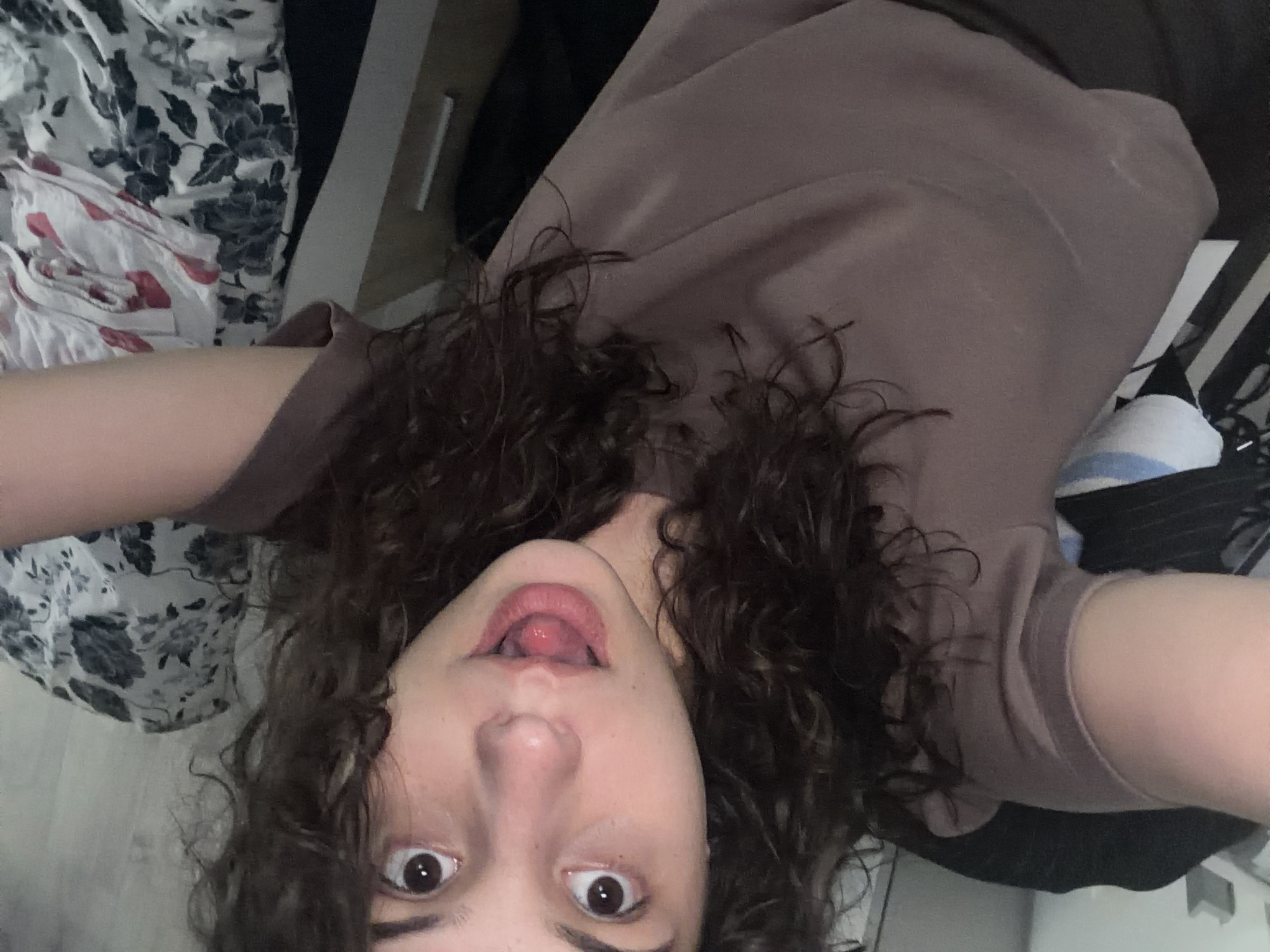 A young female with dark, curly hair is shown in a selfie-style photograph, with her face tilted towards the camera and her tongue slightly sticking out. She is wearing a mauve-colored t-shirt. The background is blurred, but appears to be a bedroom setting, with a dresser and what looks like a bed with floral-patterned bedding visible. The image is taken indoors, with no indication of a specific city or location.