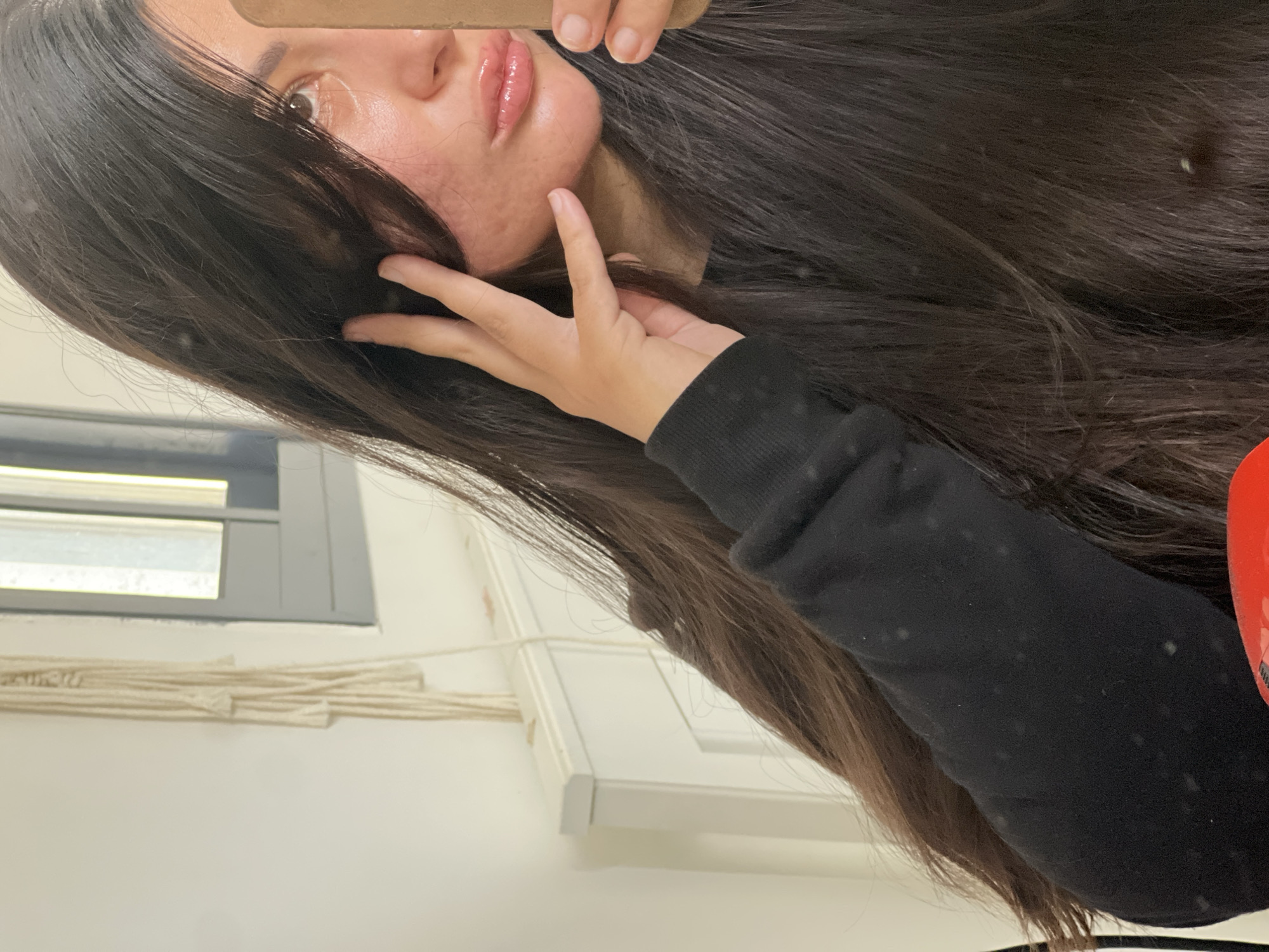 A selfie-style photo shows a person with long, dark hair partially obscuring their face. The visible portion of their face includes a portion of their eye, nose, and lips, which appear to have a glossy finish. Their hand is raised, with fingers splayed near their face. They are wearing a black long-sleeved garment. The background includes a window with a gray frame, and white walls. There are grouped strands hanging down from the wall. There is no explicit information to confirm the location in the city of Kiryat Bialik, Israel.