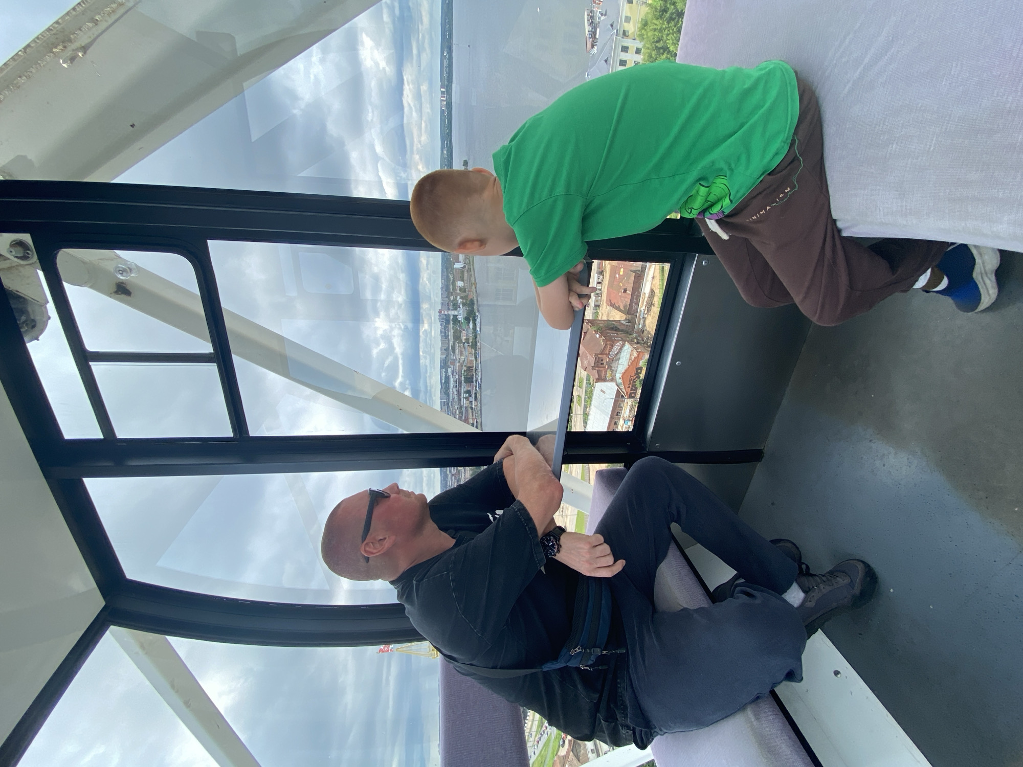 This media file shows two people inside a gondola, likely from a Ferris wheel, with a view of a city and a body of water through the windows.
The subjects are an adult male and a young boy. The man is sitting on a bench, wearing a black t-shirt, dark pants, sunglasses, and a watch. He is looking upwards and to his left, with his arms crossed. A small blue bag is strapped across his chest. The boy is standing, leaning over a railing and looking out the window. He is wearing a bright green t-shirt and brown sweatpants. He is leaning on his forearms on the railing.
The scene is a city with buildings and a river or lake visible in the background. The sky is cloudy, suggesting it is daytime. The gondola appears to be made of metal with large glass windows. The overall mood seems relaxed, with the subjects enjoying the view. There is some text on the boy's sweatpants that reads "MINIMAXISM".