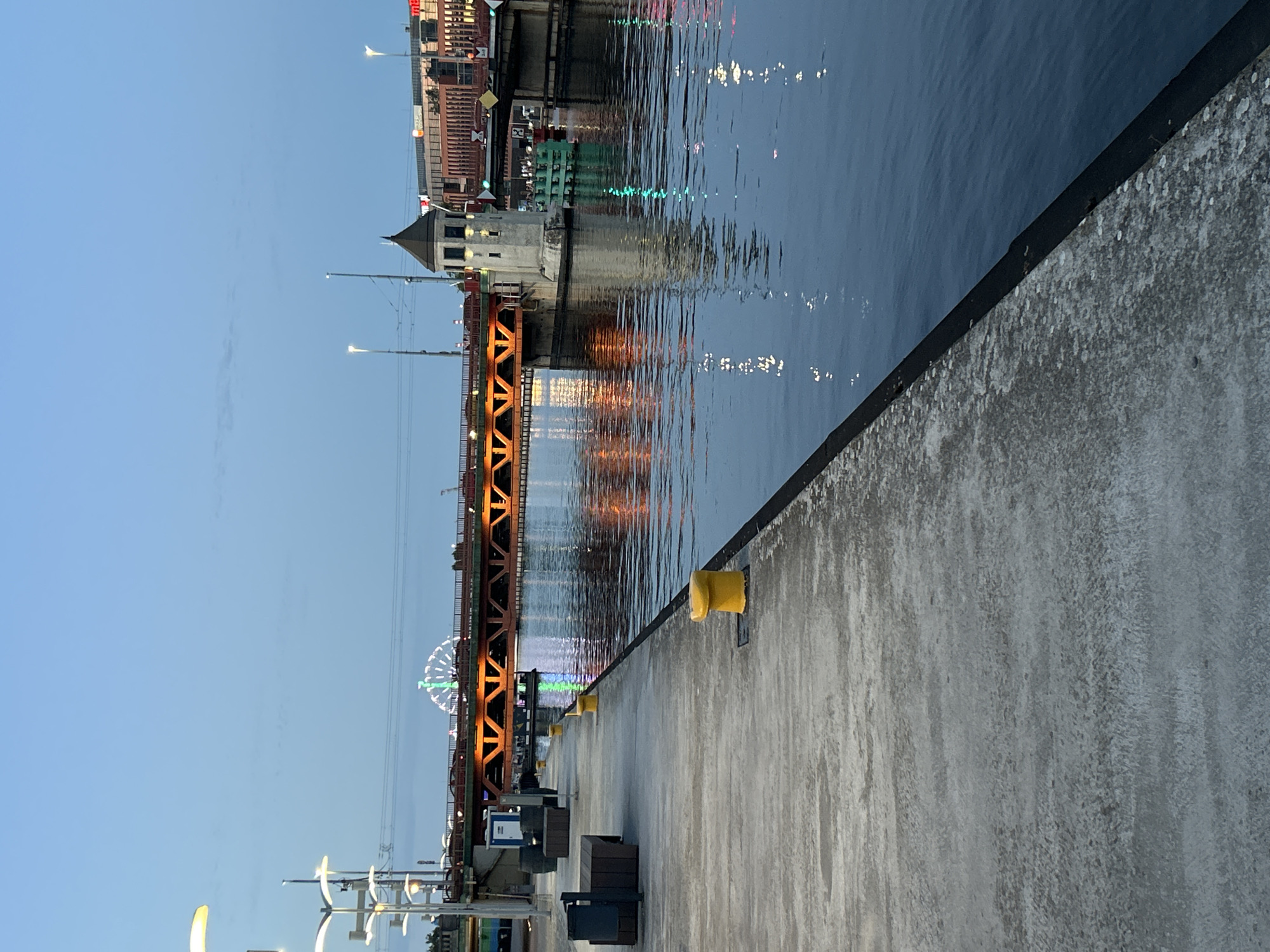 The image depicts a waterfront scene at dusk, likely in the city of Szczecin, Poland. A concrete promenade runs along the left side of the frame, bordered by dark water on the right. Several yellow bollards are visible on the promenade, and in the distance, there are benches and trash receptacles. A distinctive red bridge spans the water, illuminated by warm orange lights. Beyond the bridge, a Ferris wheel with green lights is visible, suggesting a possible fairground or entertainment area. On the right side of the water, a stone tower with a dark conical roof stands, and behind it, buildings with illuminated windows rise. The water reflects the lights from the bridge and the buildings, creating a shimmering effect. The sky is a clear, pale blue, transitioning from twilight. There are no people or animals clearly visible in the image, and no overt actions or interactions are occurring. The scene is static and serene.