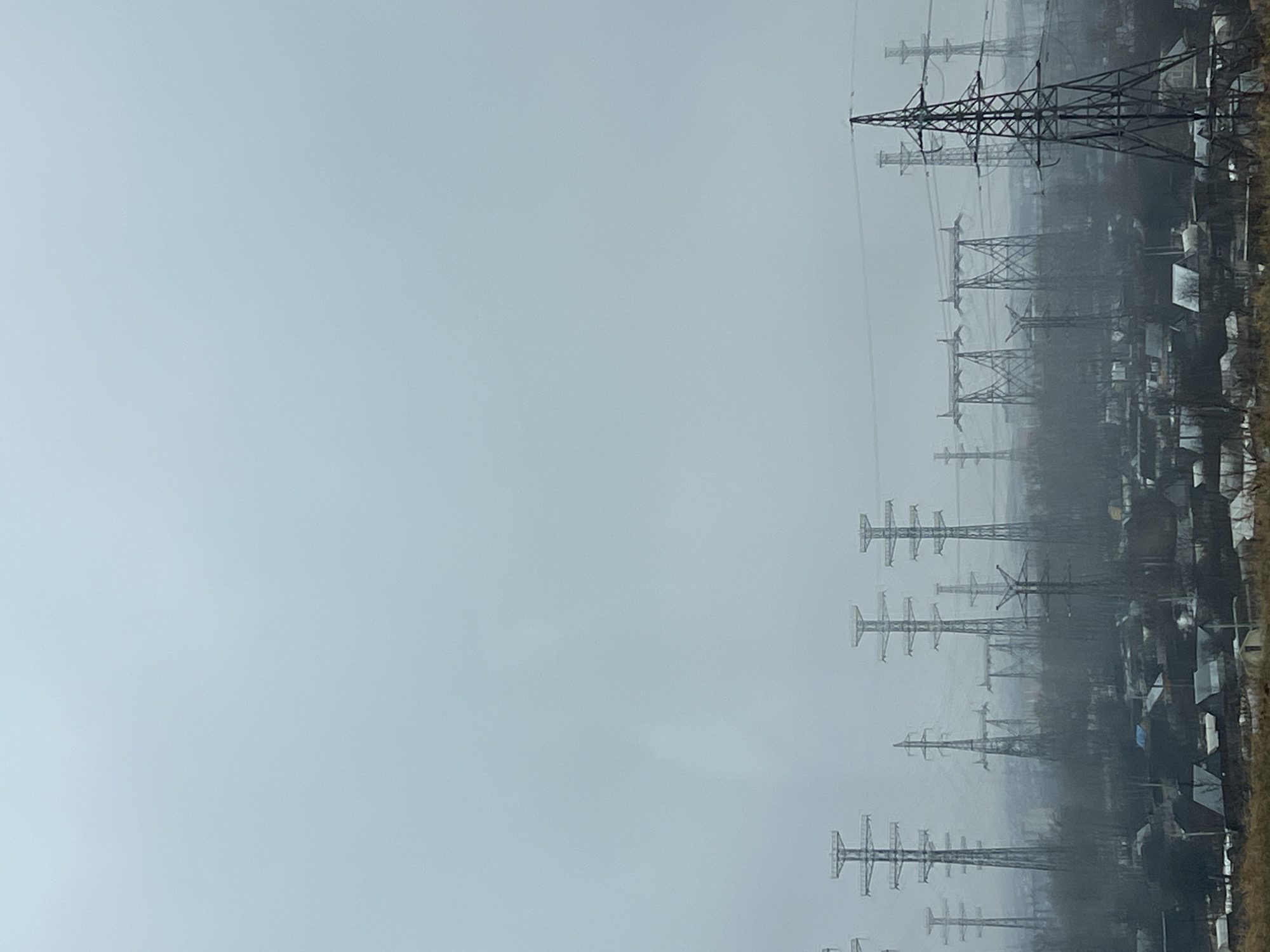 The image depicts a landscape with multiple electrical transmission towers silhouetted against a gray, overcast sky. The towers appear to be situated near or within a residential area, with rooftops visible interspersed between the towers. The scene is foggy or hazy, obscuring details and depth. The location is Chaikovsky, Russia.