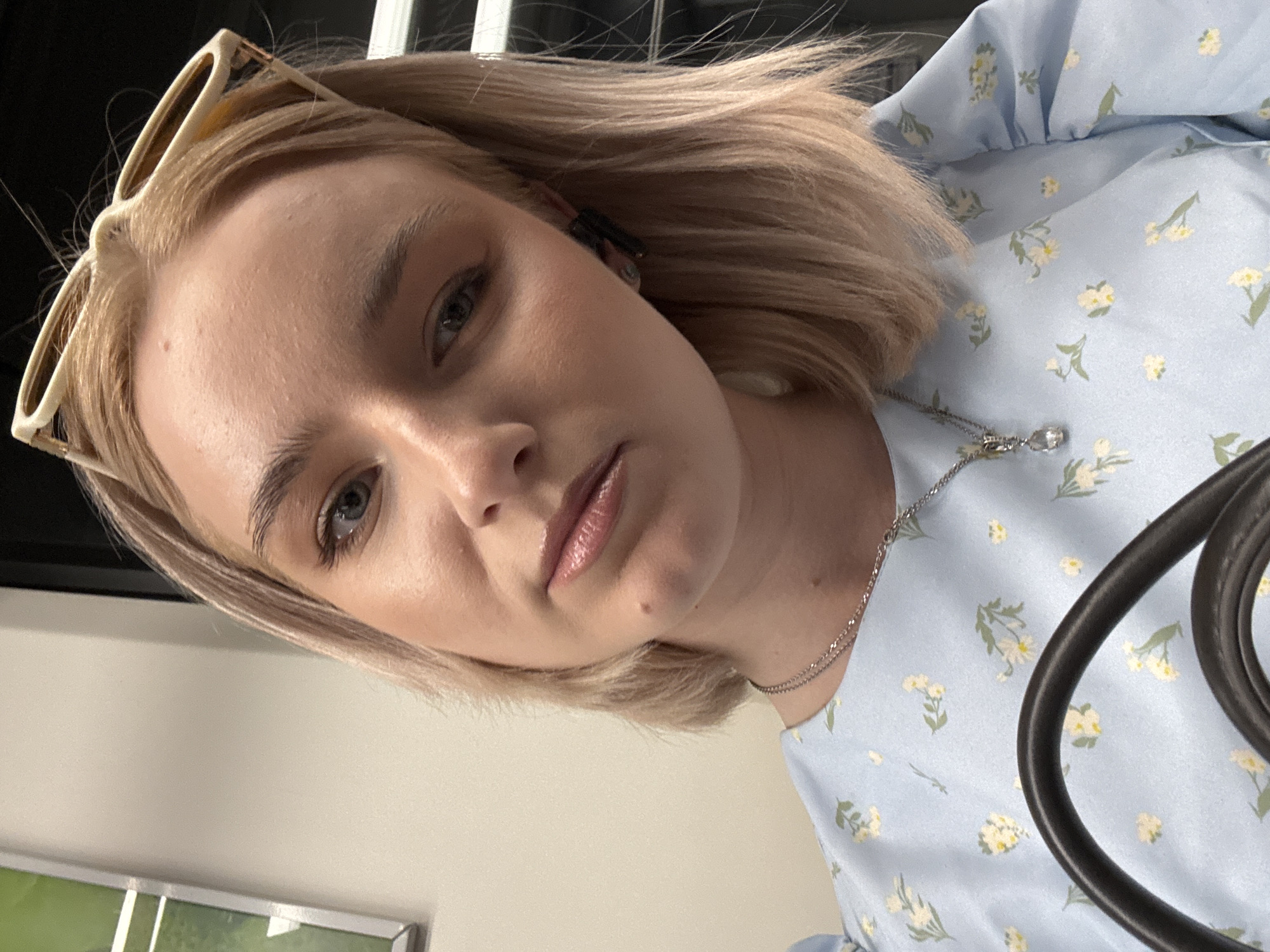 A close-up, slightly upward-angled selfie depicts a young adult female with shoulder-length blonde hair, light blue eyes, and a neutral expression. She wears a light blue top adorned with a pattern of small white and yellow flowers with green leaves. A silver-toned necklace with a clear teardrop pendant rests on her chest. Cream-colored sunglasses with dark lenses are pushed back onto her head, and a dark earbud is visible in her right ear. Her complexion is fair, with a small mole visible on her jawline. The background features a plain light beige wall on the left, with a framed green object partially visible. The upper right background is dark, suggesting an interior setting, possibly within public transport. This scene is situated in the city of Saint Petersburg, Russia.