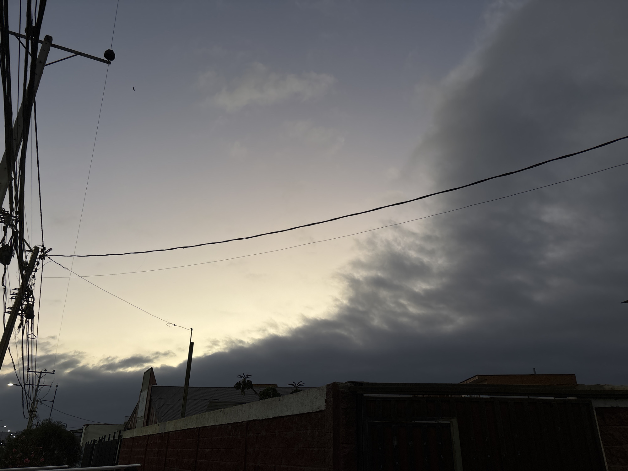 The image shows a street scene in Arica, Chile, at dusk or dawn. Overhead, a sky is filled with a mix of light gray and dark gray clouds. An electrical pole with wires dominates the left side of the frame, and additional utility wires stretch across the sky. In the lower part of the image are buildings and a brick wall bordering a property. A streetlight is visible in the distance, indicating a residential or urban area. A small object, possibly a bird, is silhouetted against the light part of the sky.
