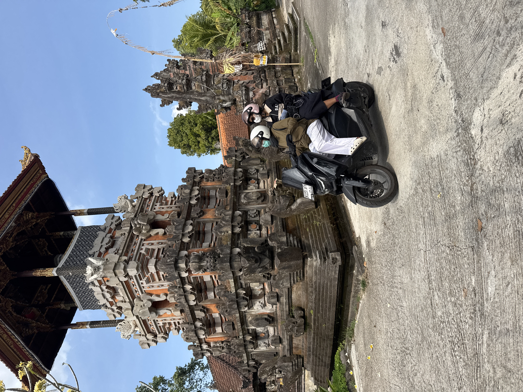 Two individuals, wearing helmets and face masks, ride on a black scooter. The person in front wears a brown jacket and white pants, while the person behind wears a black top with "GA" printed on it and dark pants. They are positioned on a paved pathway in front of a traditional Balinese structure in Gianyar, Indonesia. The structure is ornate, with intricate stone carvings and multiple tiers. In the background, trees and more traditional architecture are visible under a clear blue sky. A tall, thin pole with decorative elements stands to the right of the main structure.