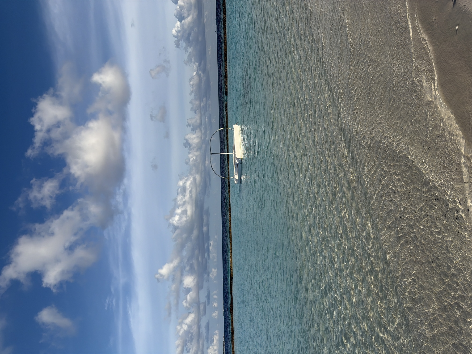 The image depicts a serene seascape in Halaveli, Maldives. The foreground shows a sandy beach meeting clear, shallow turquoise water, with sunlight creating intricate patterns on the seabed. In the mid-ground, a small, white floating platform with a curved metal frame is anchored in the tranquil ocean. The water extends to a dark blue horizon line, beyond which lies a faint, darker line of distant land or structures, possibly another island or resort. The sky above is a vibrant blue, dotted with fluffy white and grey clouds, suggesting a pleasant day. The overall scene is one of peacefulness and natural beauty.