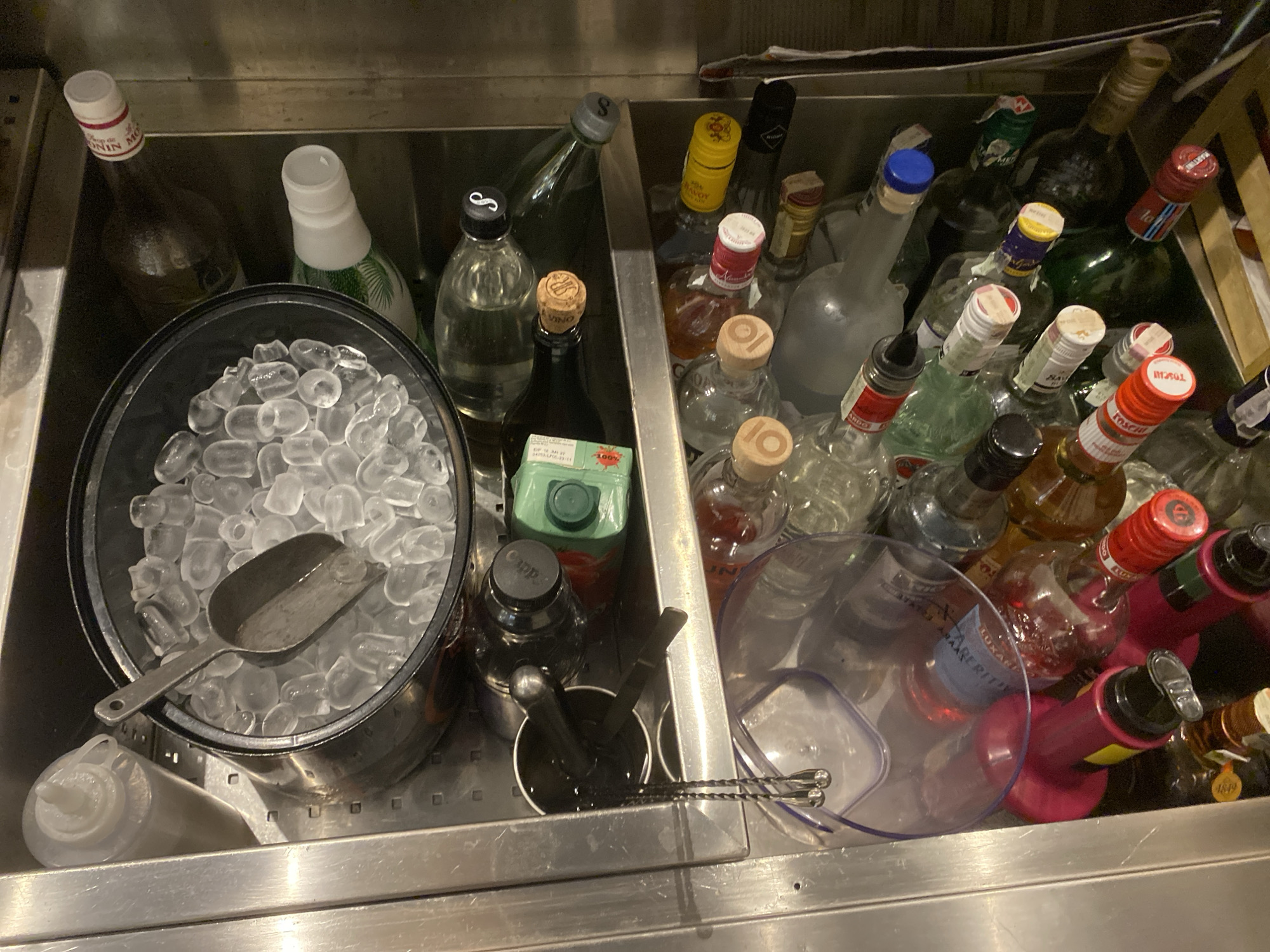 The image shows a stainless steel bar sink, divided into two compartments. The left compartment contains a large metal ice bucket filled with ice and a scoop, along with a bottle of Monin syrup, a bottle with a palm tree design, a black bottle with a "Cappa" label, a container with bar tools, and other bottles. The right compartment is densely packed with various liquor bottles, some partially obscured by clear plastic pitchers. The location in Varna, Bulgaria, is not discernible from the image. The scene appears to depict a bar's workstation, organized for drink preparation.