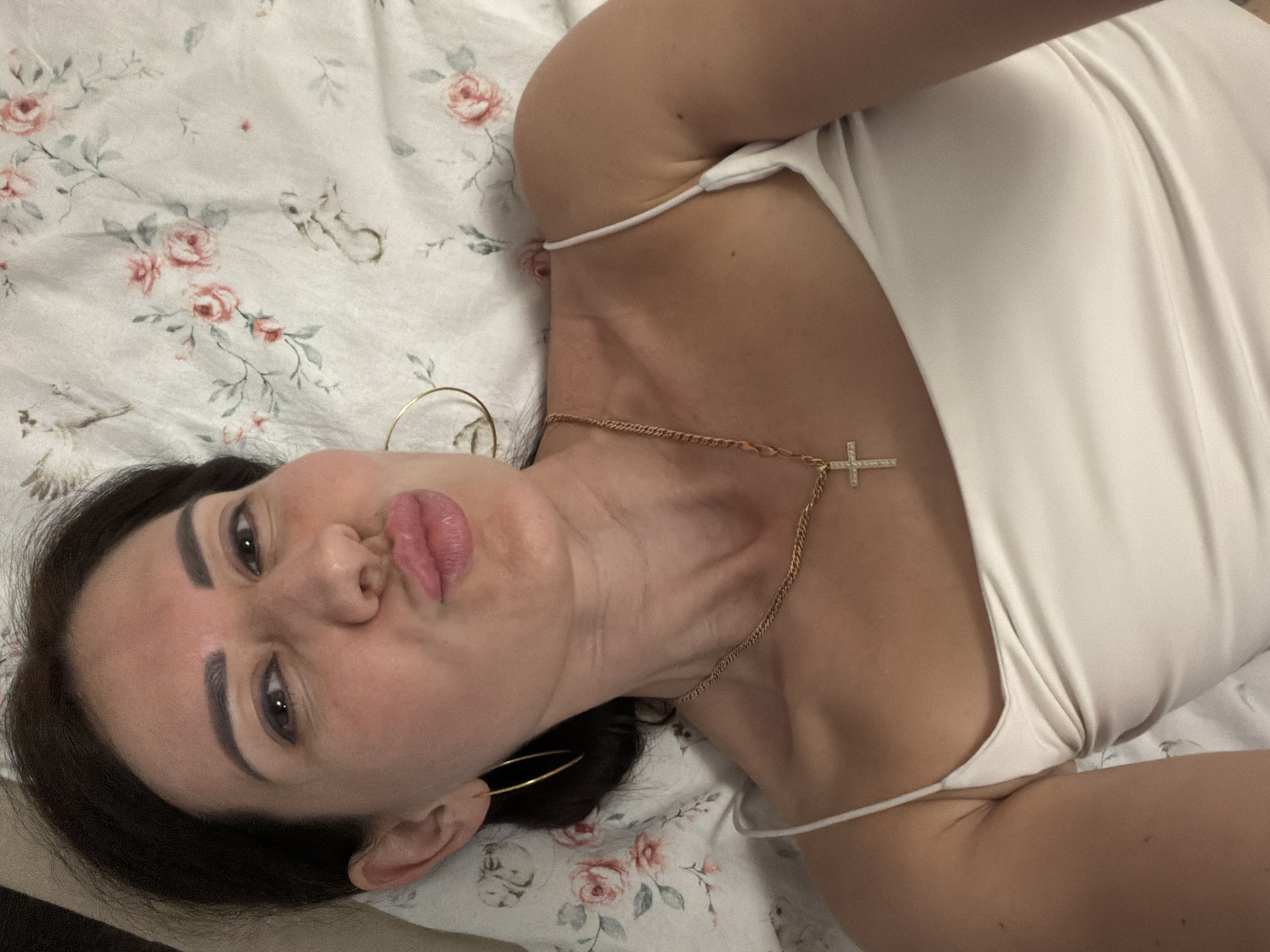 A woman with dark hair is lying on her side on a white bedspread with a floral pattern. She is wearing a white tank top and gold hoop earrings. A gold chain with a cross pendant rests on her décolletage. Her lips are pursed as if she is about to kiss. The background is a close-up of the floral bedspread, which features pink roses and green leaves, as well as small illustrations of birds and rabbits. The image appears to be a selfie taken indoors, possibly in a bedroom. The location in Sukhodol, Russia is not evident from the image.