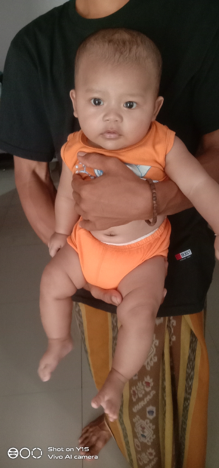 An indoor scene in Brumbungan Kidul, Indonesia, depicts an adult holding an infant. The adult, partially visible from the chest down, wears a dark-colored short-sleeved shirt with a white "B3ST" logo on the lower right and a yellow and brown patterned sarong. A brown beaded bracelet is present on the adult's right wrist. The infant, with a light complexion and sparse dark hair, is dressed in an orange sleeveless top with faint white graphics and matching orange diaper cover-shorts, revealing the navel. The infant is held upright, looking forward with a neutral expression. The adult's bare feet are visible on a light gray tiled floor. The background is indistinct.