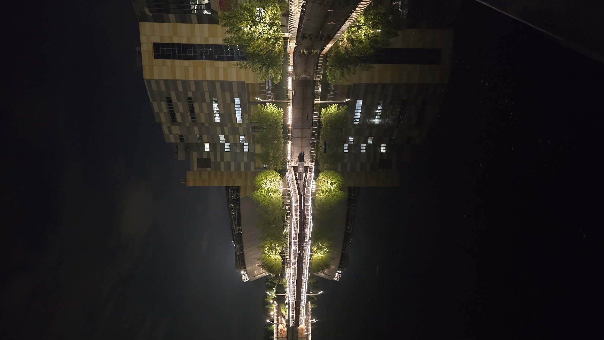 The image depicts an urban night scene captured across a calm body of water, providing clear reflections of architectural structures and illuminated foliage. On the right, a large, multi-story modern building with a distinctive facade featuring a checkerboard pattern of light grey, dark grey, and beige/yellow panels is prominently visible. Numerous rectangular windows on this building are illuminated, indicating interior lighting. To its left, a large, angular structure with a curved, overhanging roof and a light-colored, possibly metallic exterior, suggestive of an arena or event complex, is softly illuminated. Along the near bank of the dark water body, a promenade or walkway is lined with evenly spaced warm yellow lights. These lights also illuminate several mature trees, highlighting their vibrant green foliage. Railings are visible along the promenade edge. The still surface of the water perfectly mirrors the buildings, illuminated trees, and linear promenade lighting, creating a symmetrical reflection. The sky above is dark, with faint cloud formations discernible. No individuals or explicit actions are visible.
This scene is located in Kyiv, Ukraine, specifically along the **Pochayna River/Canal**. The prominent building on the right is identified as the **Pochayna Event Hall**, and the large angular structure on the left is the **Ice Arena**, both situated adjacent to the illuminated embankment of the Pochayna River/Canal.