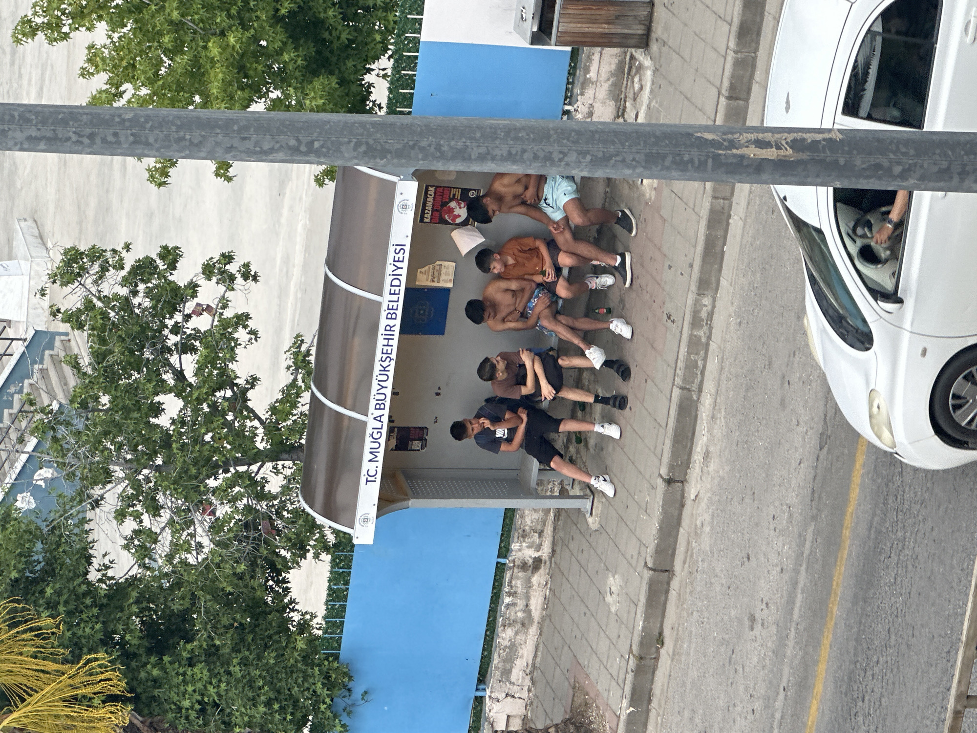 The image shows a bus stop in Fethiye, Turkey, with six young men sitting inside. The bus stop is painted blue and has the text "T.C. MUĞLA BÜYÜKŞEHİR BELEDİYESİ" written on it. The men are dressed in casual clothing, and some are shirtless. A white car is parked nearby. The scene appears to be captured from an elevated perspective.