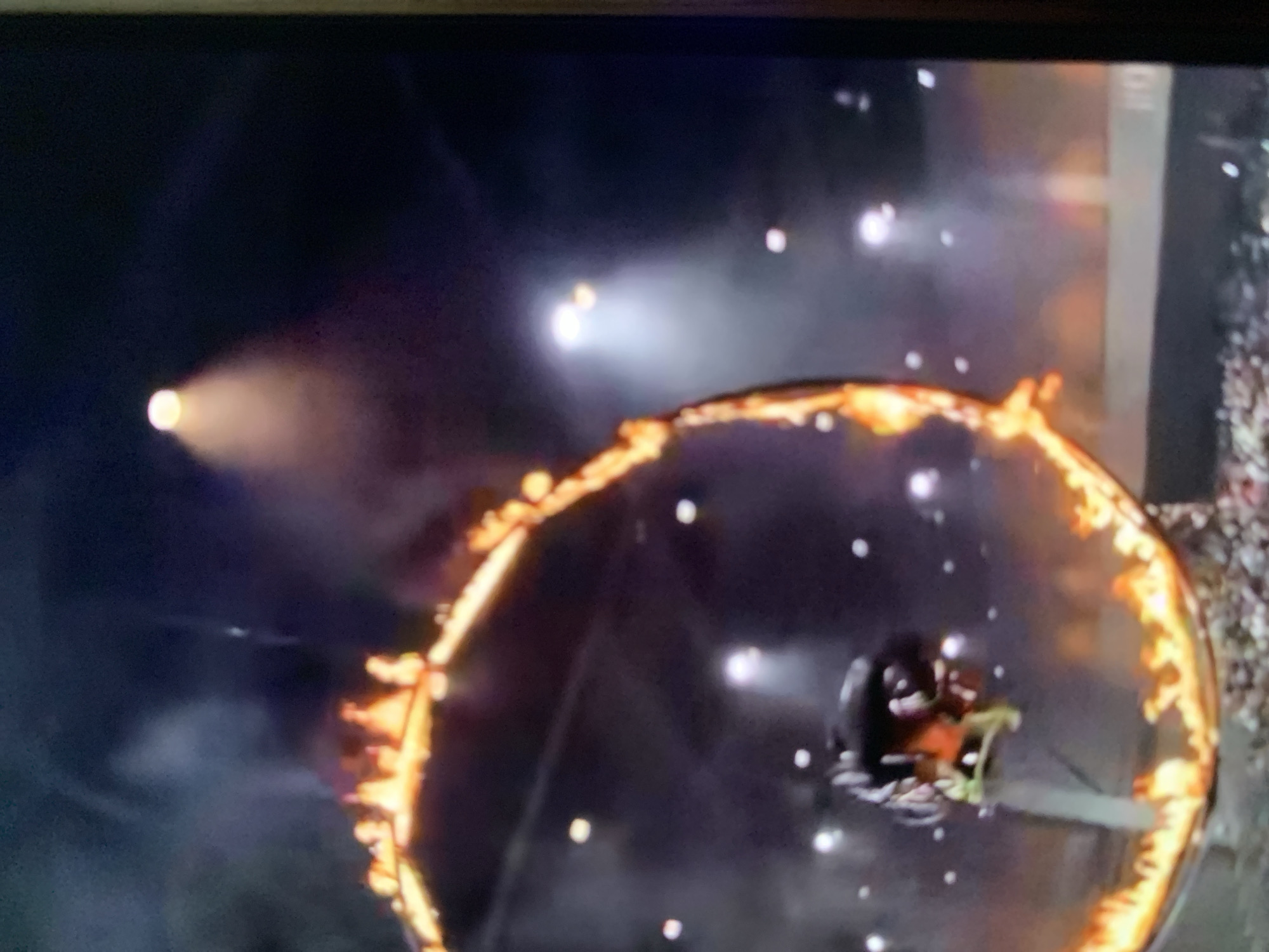 The image captures a stunt performer on a motorcycle, leaping through a flaming ring. The scene takes place in an indoor arena, evidenced by the high ceiling, spotlights, and bleachers filled with spectators. Smoke and intense flames engulf the ring, creating a dramatic and dangerous atmosphere. The performer is suspended mid-air, showcasing the daring nature of the stunt. The lighting is theatrical, with bright spotlights illuminating the action and casting shadows. The background suggests a large-scale event, possibly a circus or a motorsports show. There is no visible indication of the location being in Stavropol, Russia.