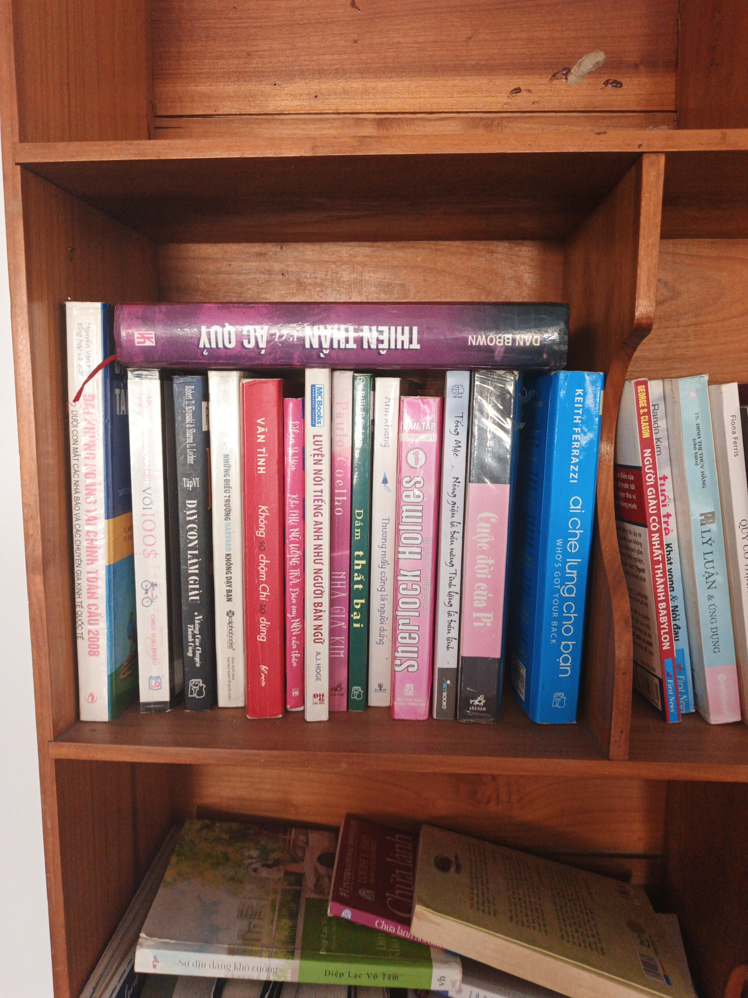 A bookshelf made of dark wood is filled with books. The top shelf has one large book with purple and black lettering that reads "THIEN THAN AC QUY" and "DAN BROWN" in English. The second shelf has a row of books with various colored spines. The books are standing upright, and some are partially obscured by others. The third shelf contains more books, some of which are stacked on top of each other. The overall impression is one of a home library or a collection of personal reading material. The environment appears to be indoors, likely a residential setting, given the presence of a bookshelf and books. The lighting is natural, suggesting daylight. There are no people visible in the image. The image is a close-up shot of the bookshelf, focusing on the books themselves and the wooden structure of the shelf.