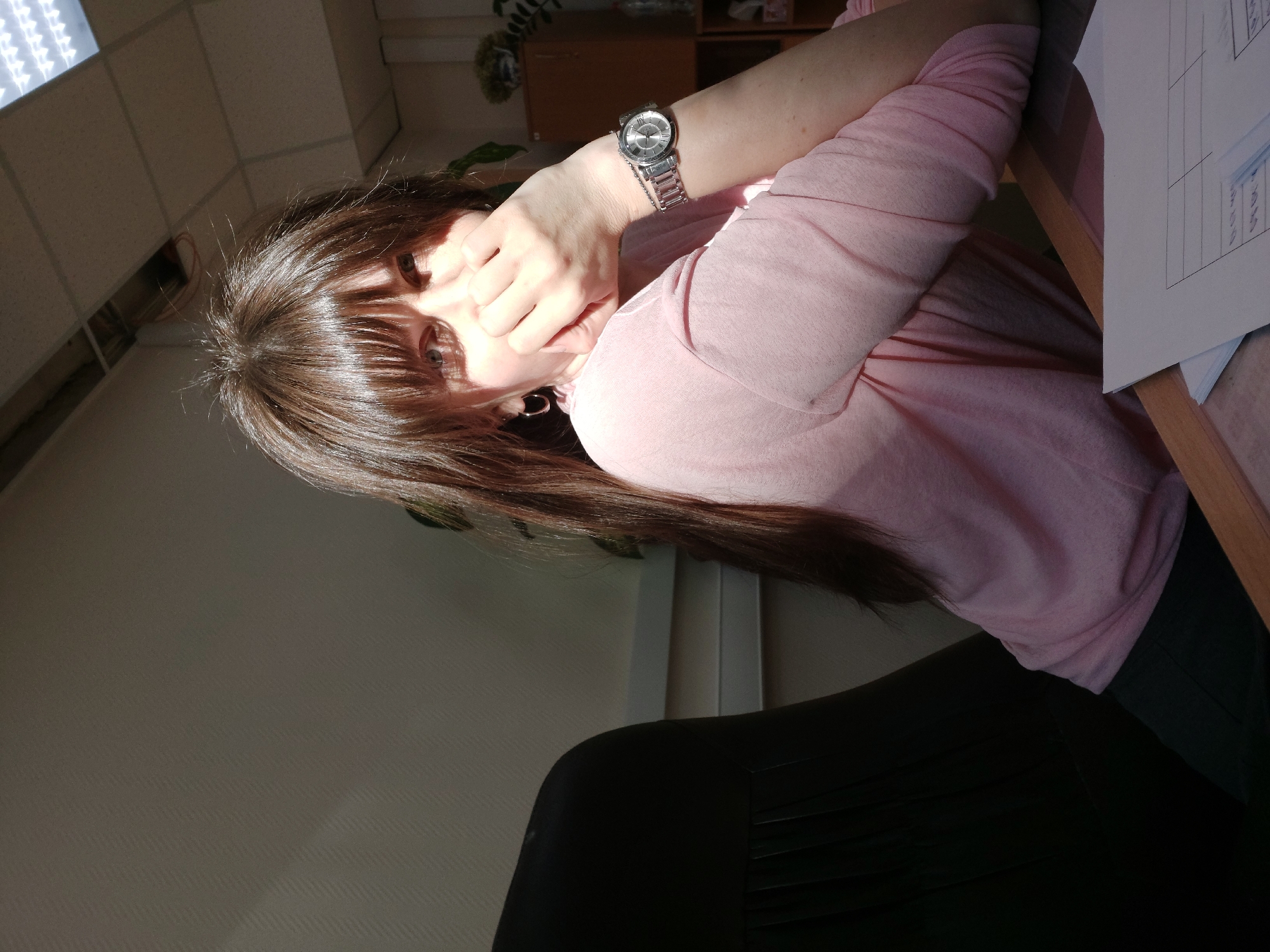 The image shows a woman with long brown hair wearing a pink shirt and a watch, sitting at a desk with papers in a room. The location is Chaikovsky, Russia. A black chair is in the foreground. The room appears to be an office with a cabinet and a light fixture visible. The woman's hand is near her face.