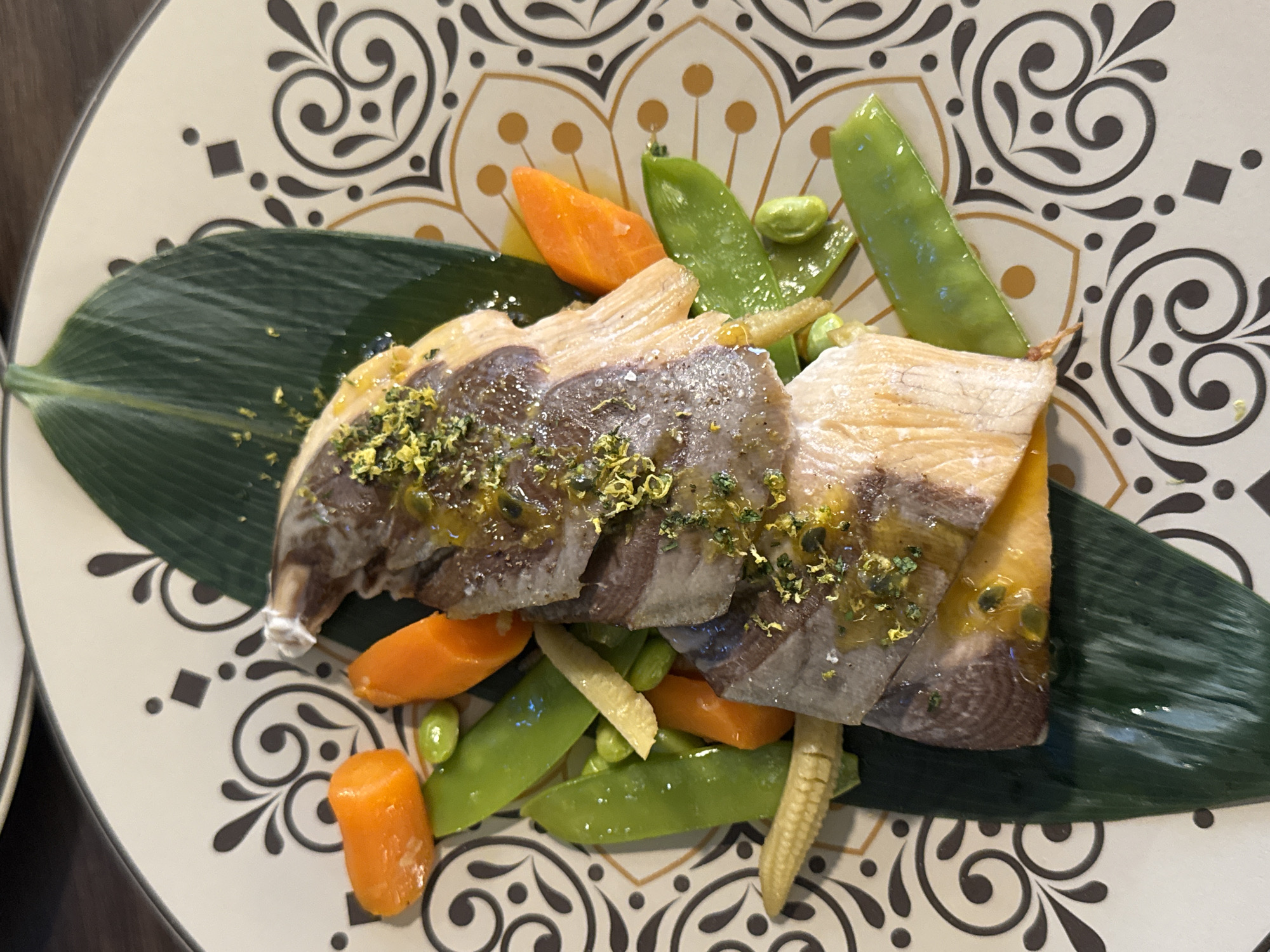A close-up, top-down shot shows a plate of food, likely salmon, with mixed vegetables. The plate has an intricate, decorative pattern with swirling black lines and small gold-colored circles on a cream background. The food is arranged on a large, dark green leaf. The salmon appears to be thinly sliced and layered, topped with a glistening sauce and what looks like finely chopped herbs or zest. Surrounding the fish are vibrant green snow peas, small orange carrot pieces, edamame beans, and tiny ears of baby corn. The lighting is bright, highlighting the textures and colors of the dish. The scene appears to be indoors, on a table surface that is dark wood. There are no people visible in the image, and the focus is entirely on the presentation of the food. There is no information about the location in Sofia, Bulgaria.